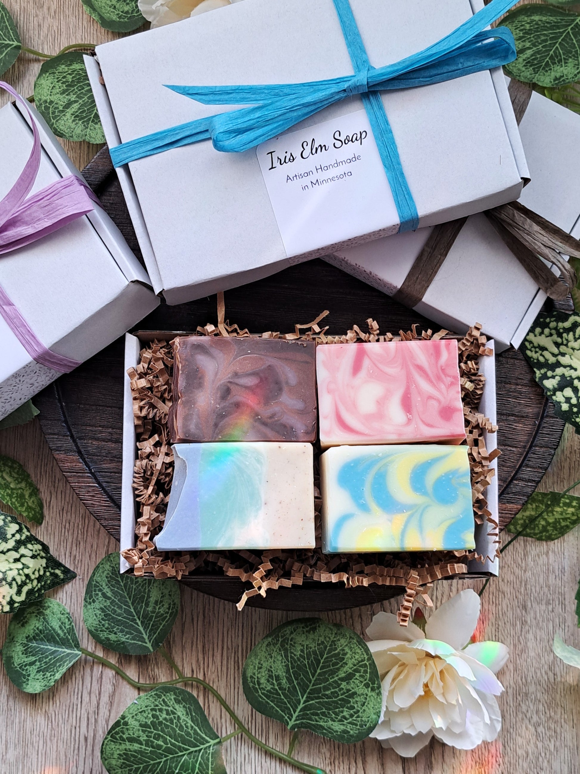 Box of colorful soap bars with a 'Iris Elm Soap' packaging on a wooden surface with green leaves.