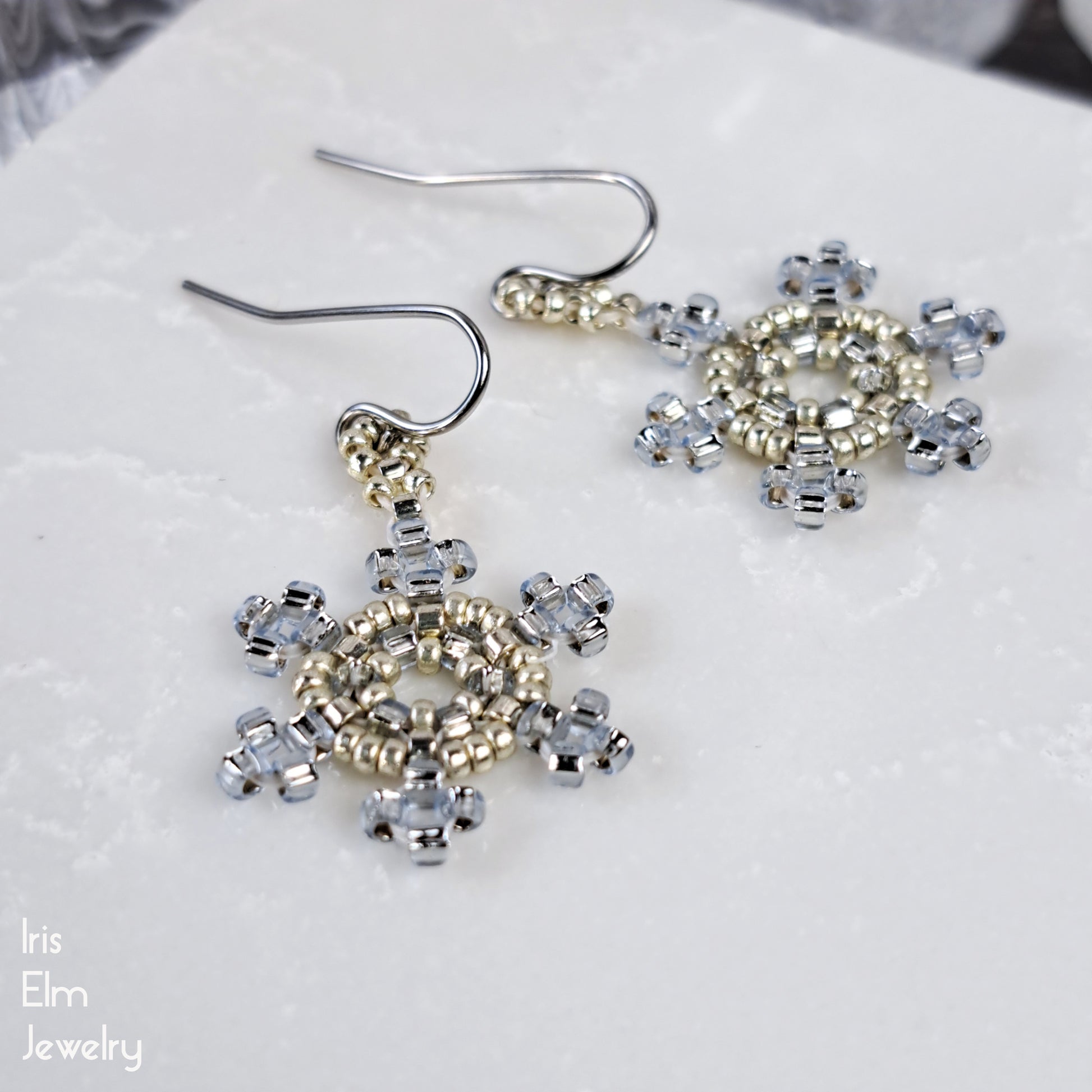 Snowflake-shaped earrings with light blue and silver beaded design on a white surface, branded 'Iris Elm Jewelry'.