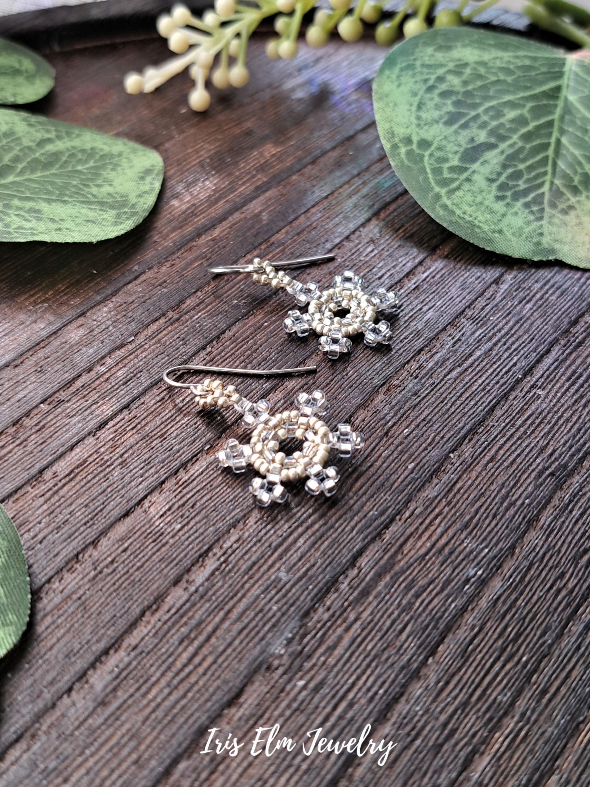 Pair of silver pale blue snowflake seed bead earrings on a wooden surface with green leaves