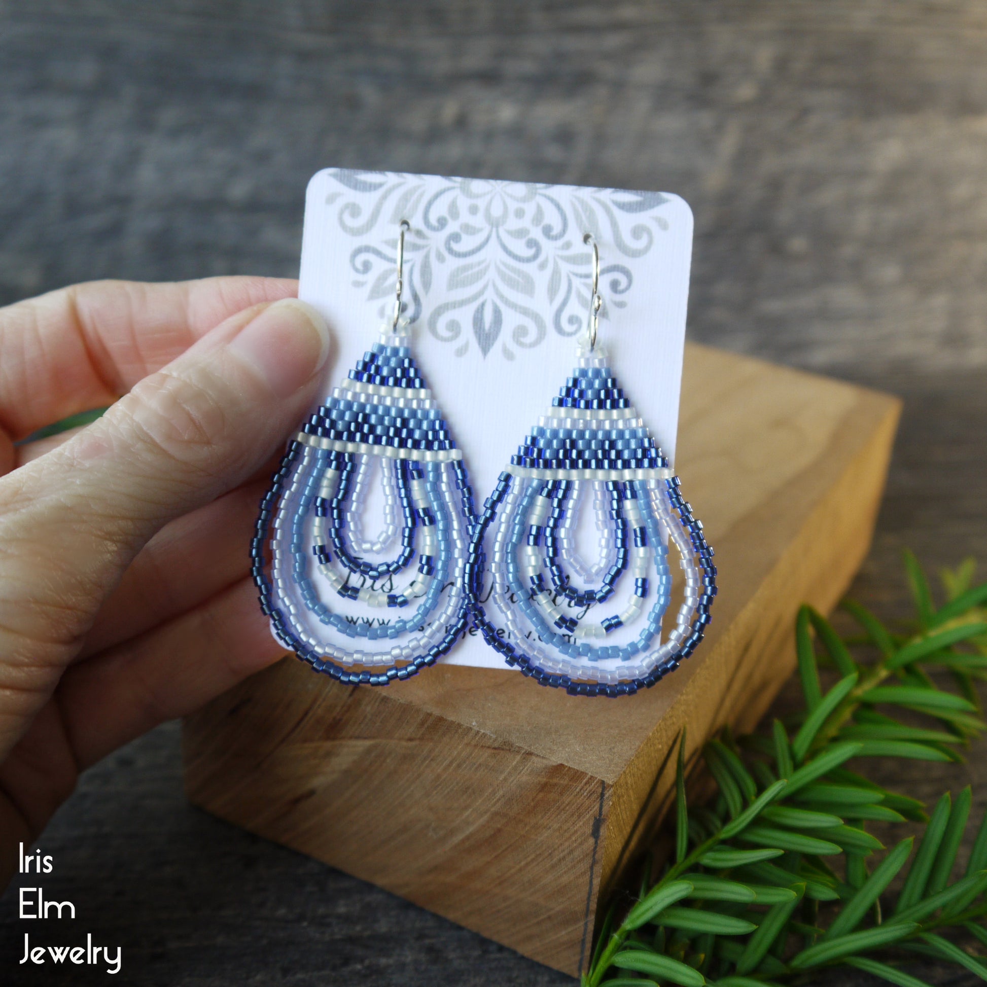 Blue beaded teardrop seed bead earrings on a card held by a hand with a wooden block and greenery in the background.