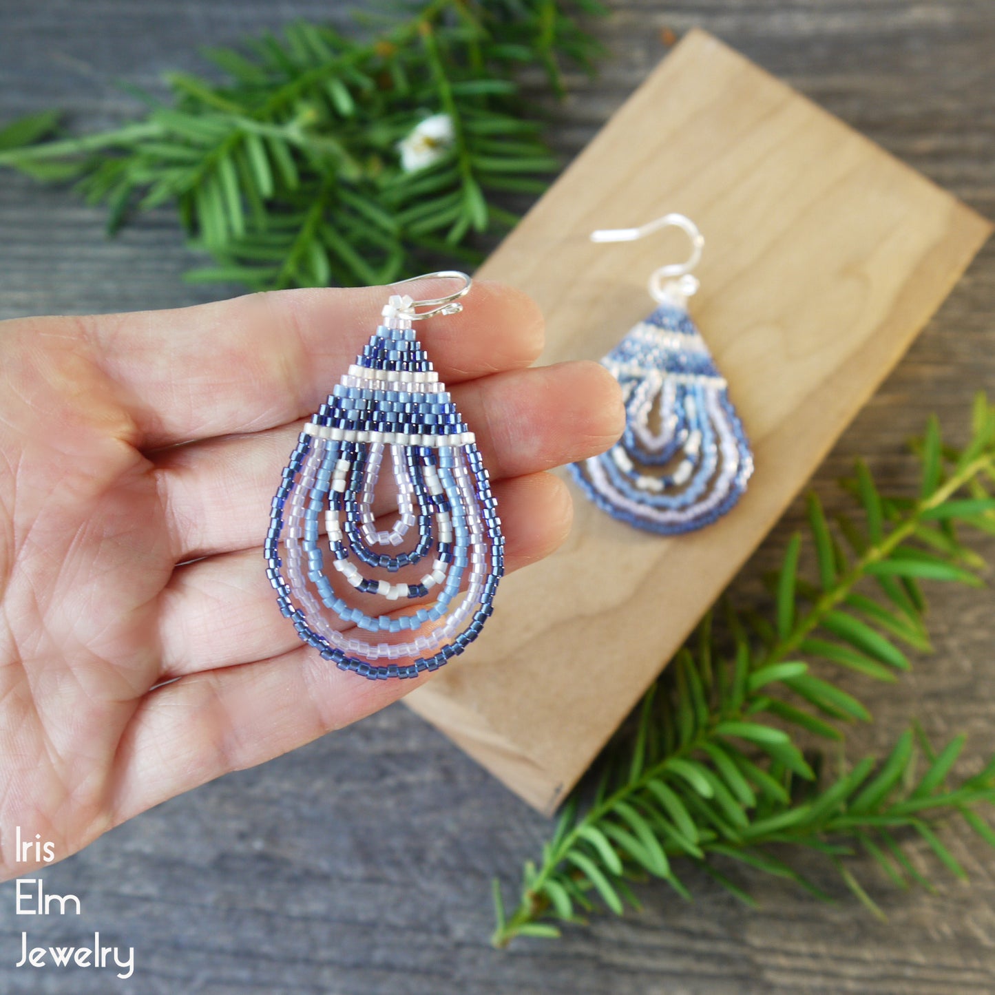Teardrop-shaped blue beaded earrings held by a hand with a wooden block and greenery in the background, featuring 'Iris Elm Jewelry' branding.
