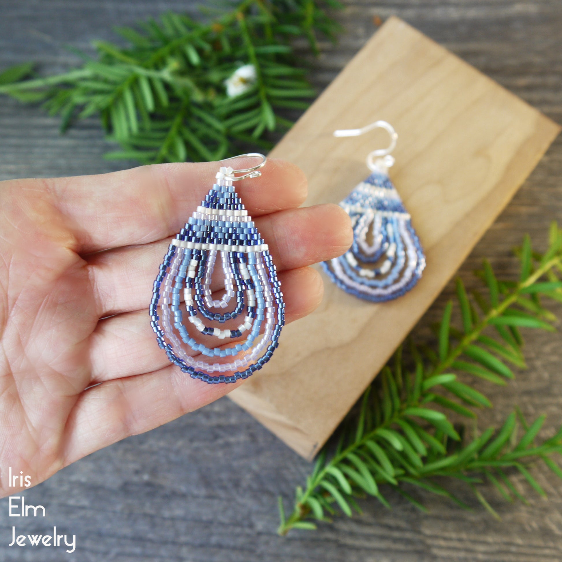 Teardrop-shaped blue beaded earrings held by a hand with a wooden block and greenery in the background, featuring 'Iris Elm Jewelry' branding.
