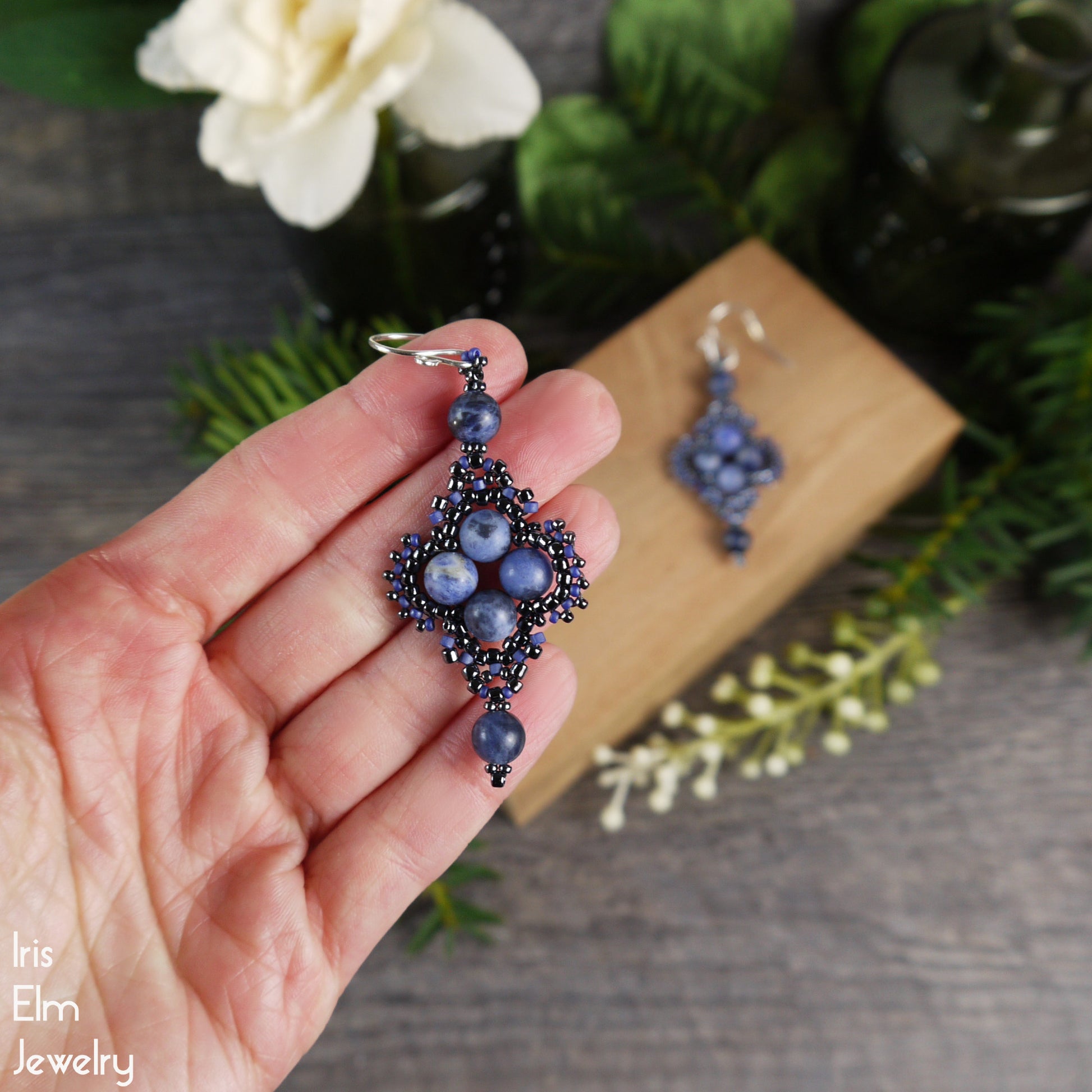 Hand holding a pair of blue beaded sodalite gemstone boho earrings with a wooden box and flowers in the background.