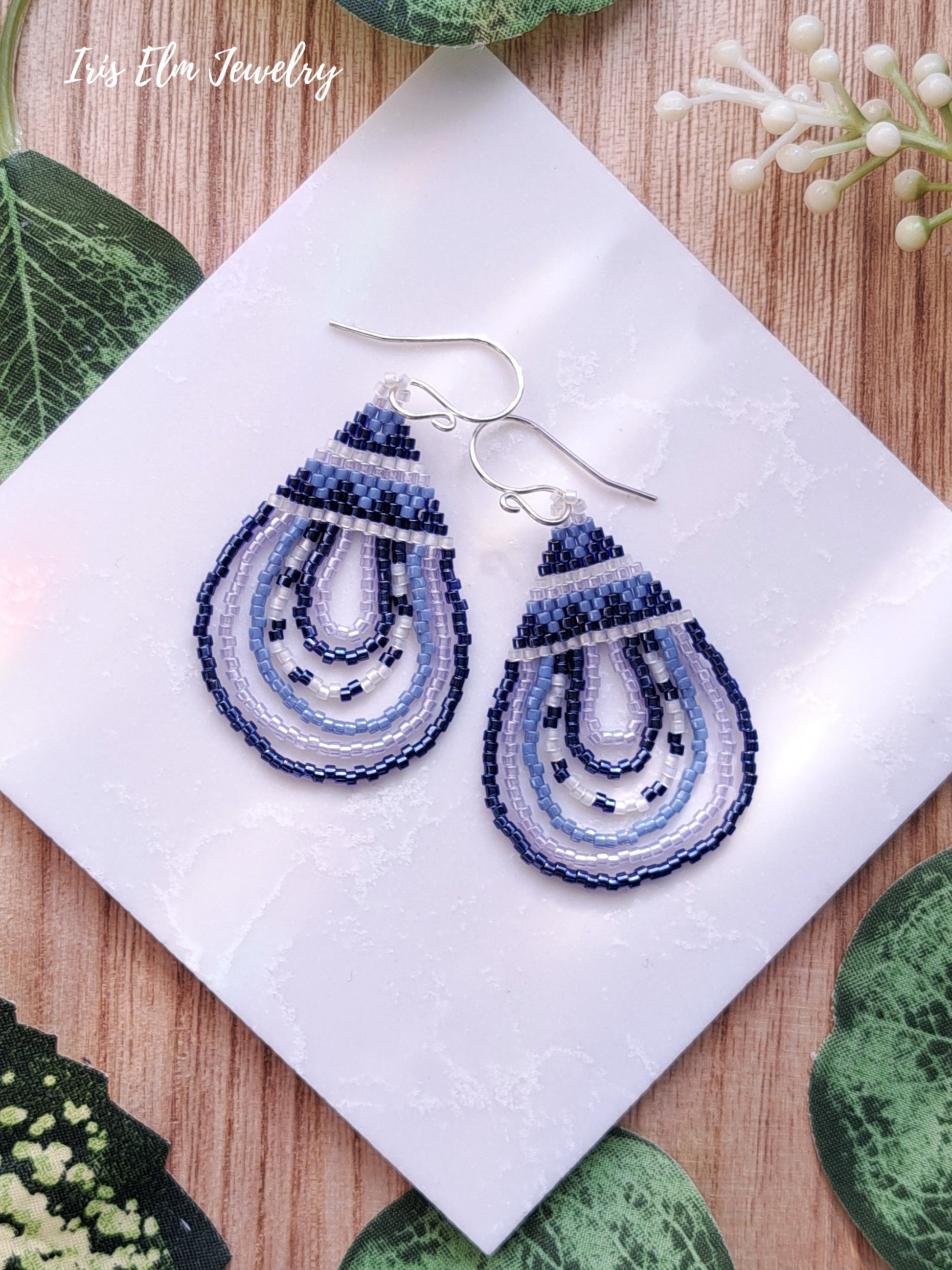 Blue beaded glass seed bead woven teardrop earrings on a white napkin with 'Iris Elm Jewelry' branding.