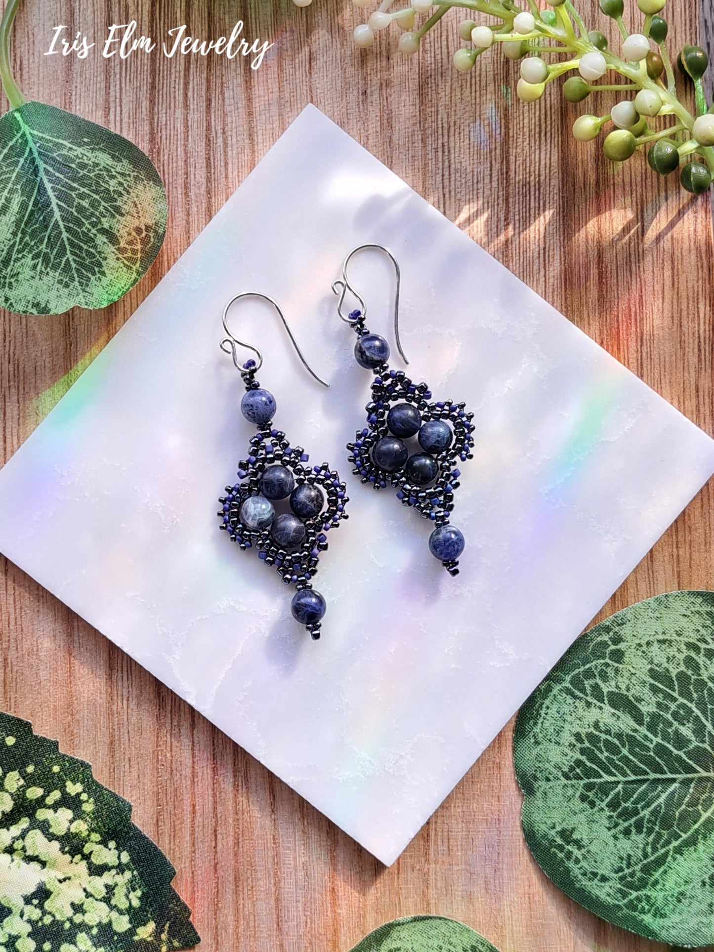 Pair of intricate blue sodalite beaded earrings on a white tile with green leaves in the background, featuring 'Iris Elm Jewelry'.