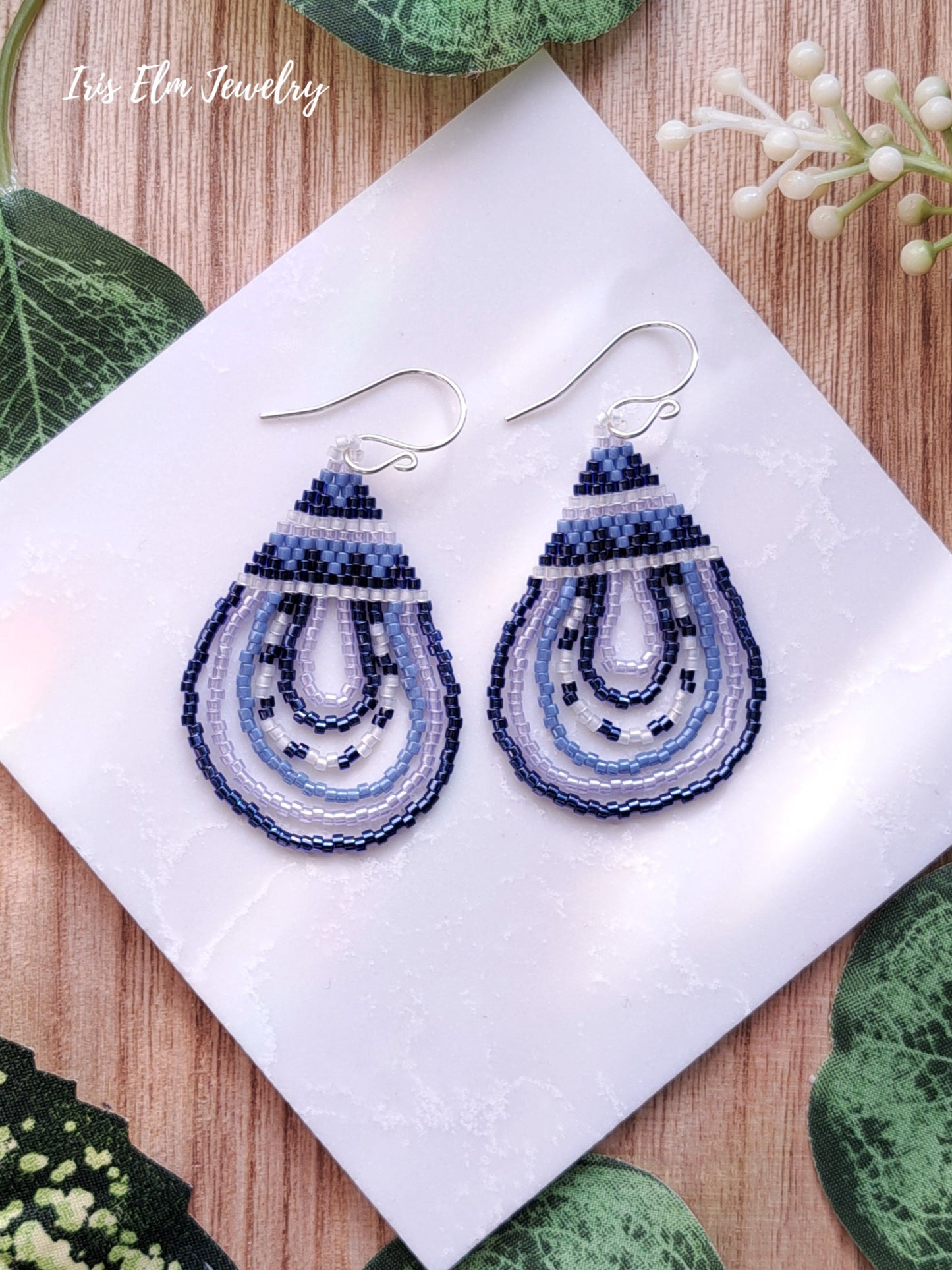 Pair of beaded blue seed bead teardrop earrings on a white cloth with green leaves in the background