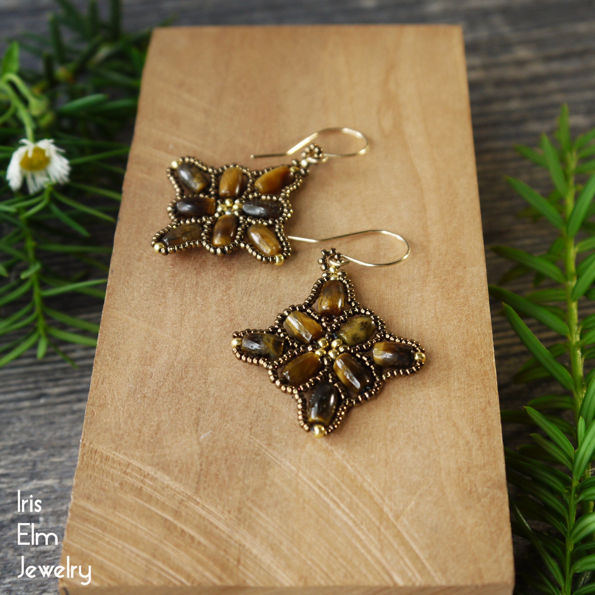 Pair of beadwoven tiger eye earrings with goldfilled ear wires on a wooden block, surrounded by greenery.
