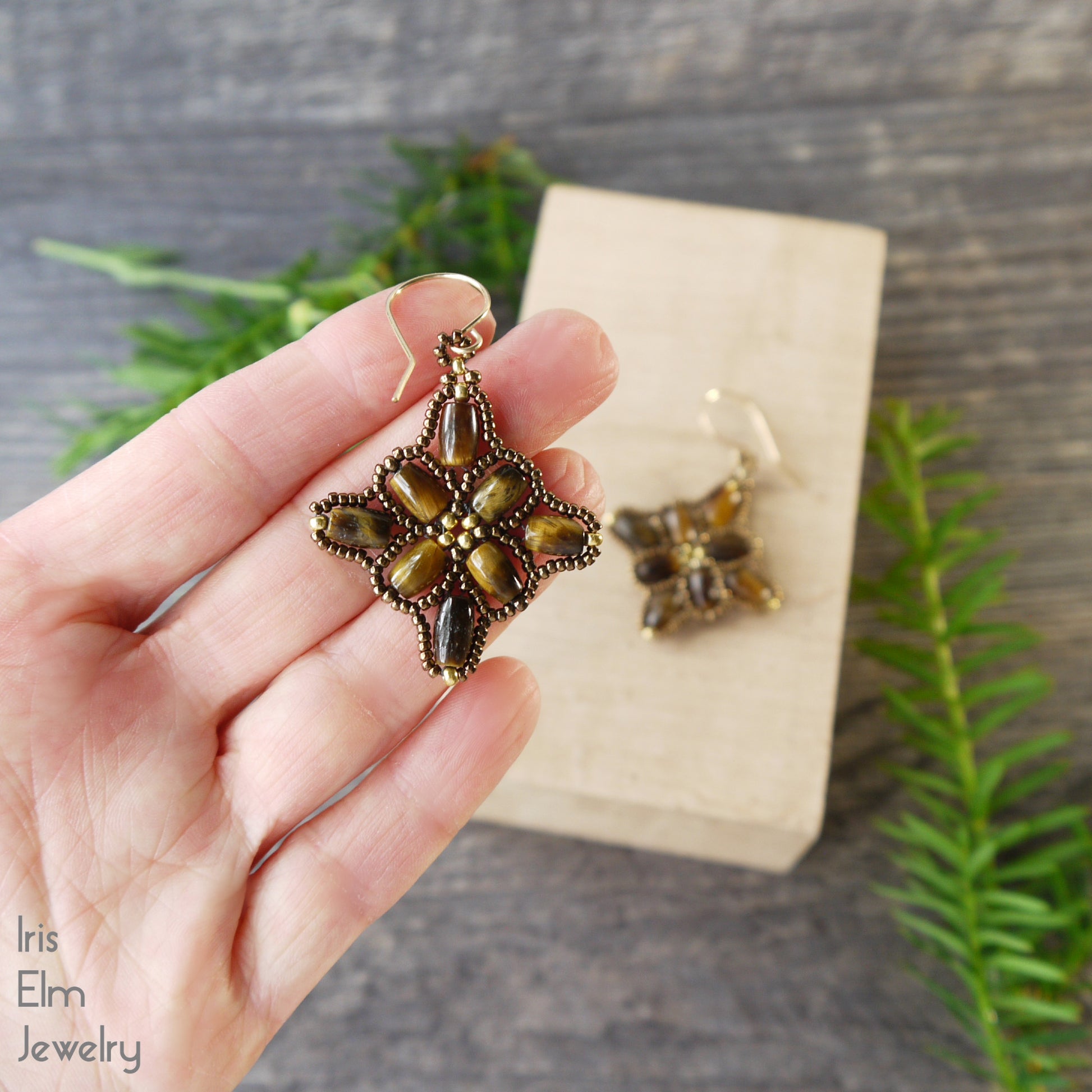 Hand holding a decorative tiger eye brown beadwork gemstone earring with a wooden block and greenery in the background, featuring 'Iris Elm Jewelry'.