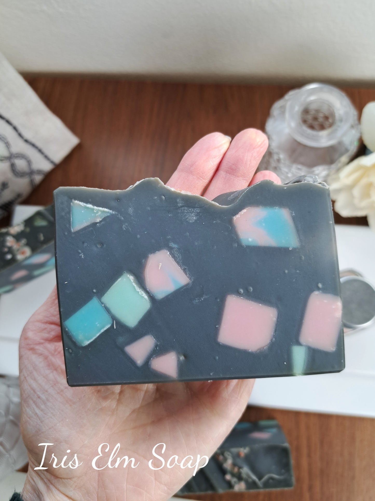 Hand holding a handmade soap bar with colorful embedded pieces on a wooden surface.