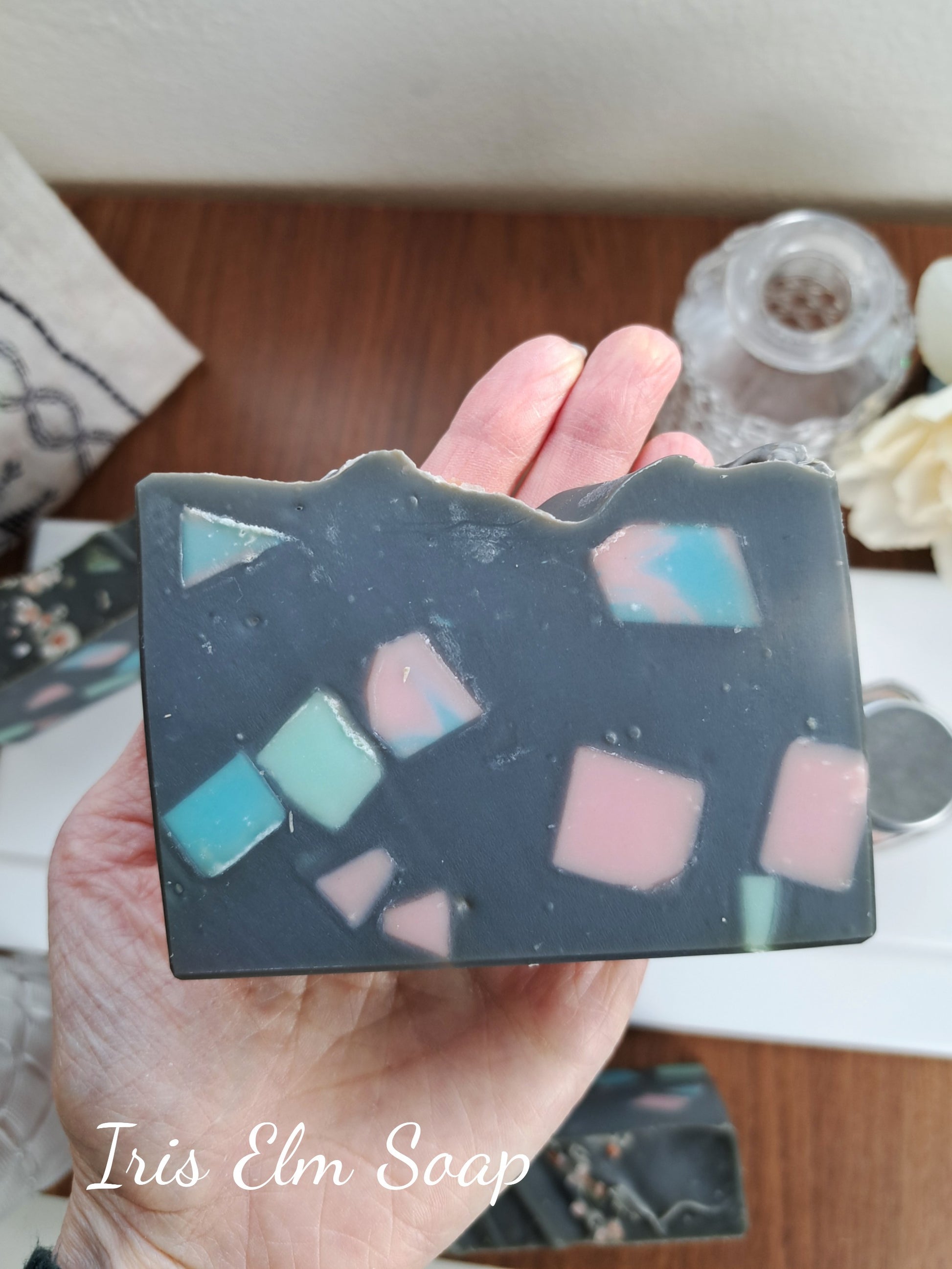 Hand holding a handmade soap bar with colorful embedded pieces on a wooden surface.