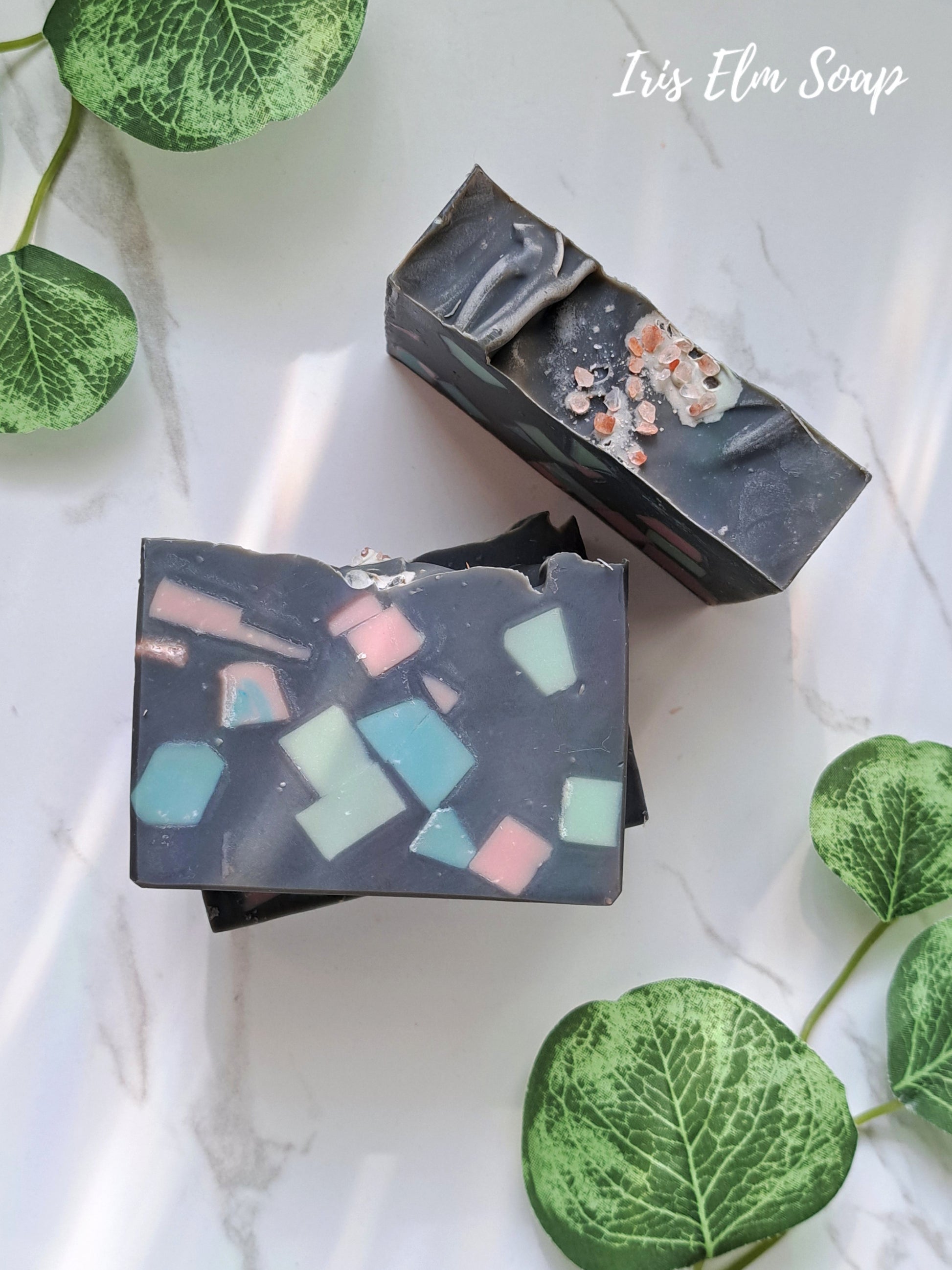 Two bars of sea salt and orchid handmade soap with a black pink sea foam green terrazzo mosaic design on a white surface with green leaves.