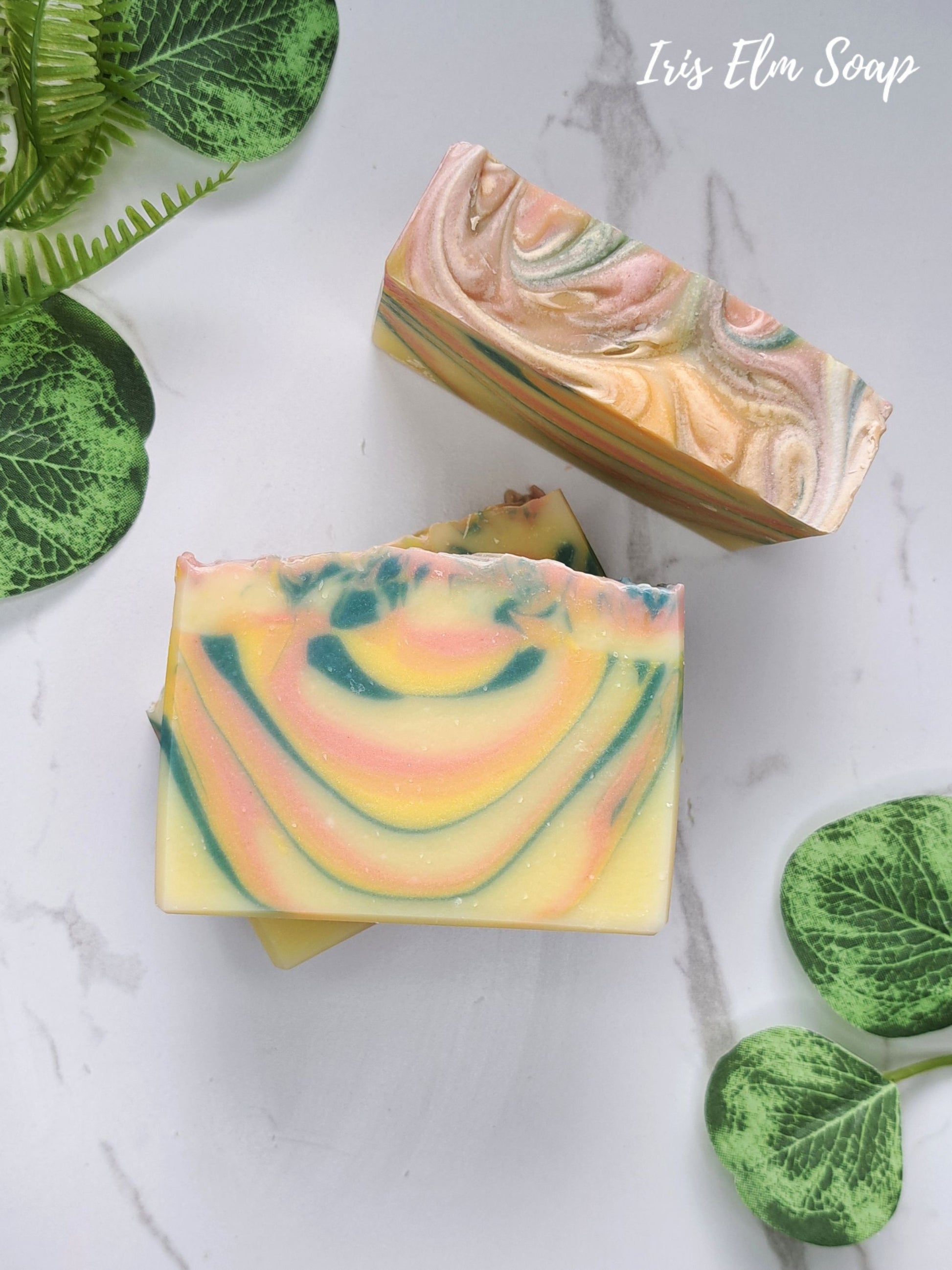 Two decorative orange patchouli handmade soap bars with marbled swirls of yellow, peach, pink, and green, arranged on a light marble surface with green plant leaves around them. The text 'Iris Elm Soap' appears in the top right corner.