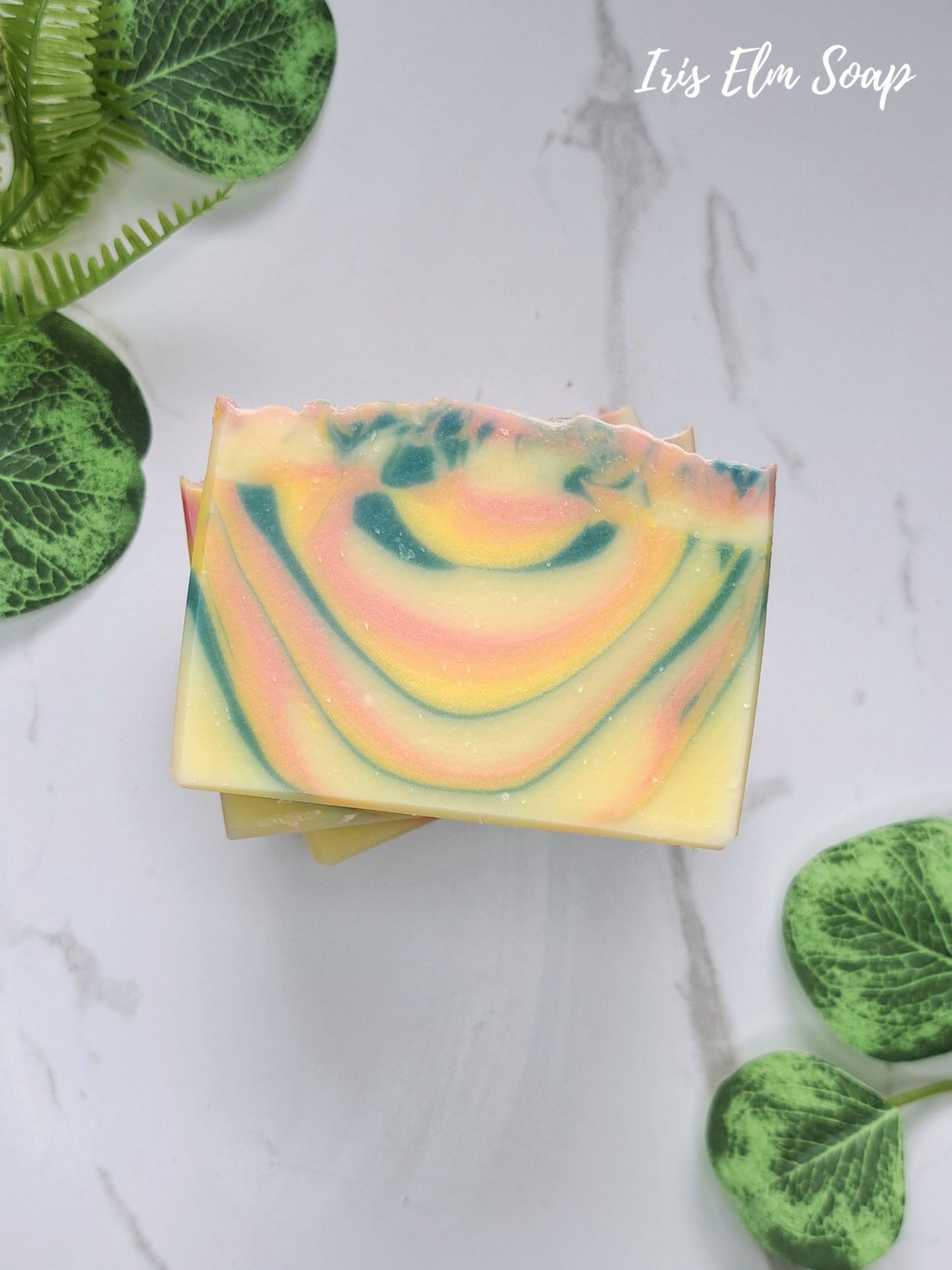 A stack of orange patchouli handmade rectangular bars of soap with a marbled swirl pattern of yellow, pink, and green, styled on a white marble background with green leaves.