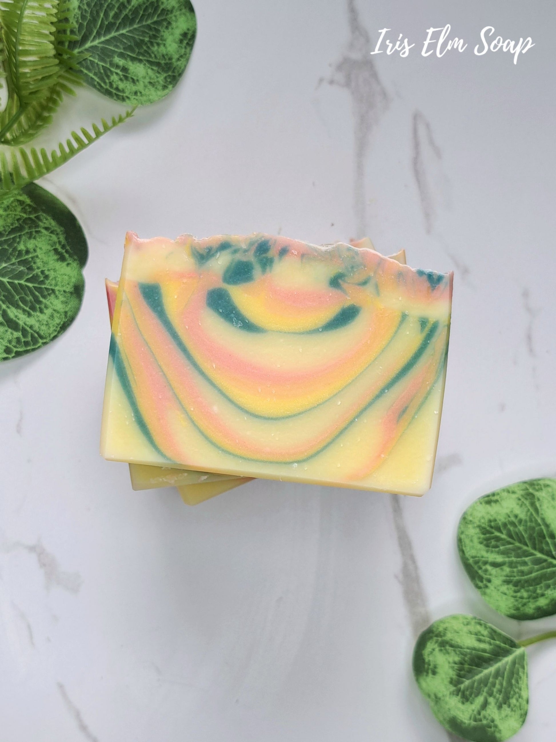 A stack of orange patchouli handmade rectangular bars of soap with a marbled swirl pattern of yellow, pink, and green, styled on a white marble background with green leaves.