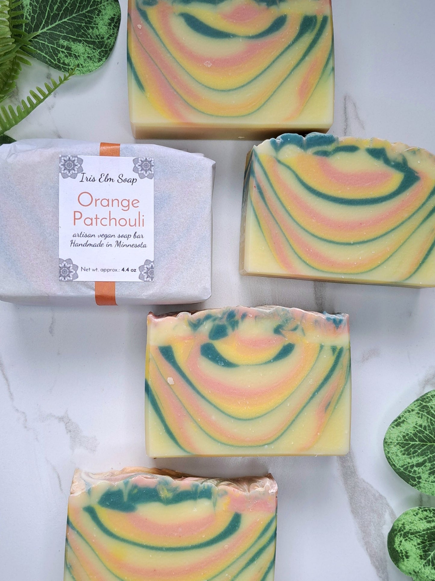 Five bars of handcrafted soap with a marbled swirl pattern of yellow, orange, and green are arranged on a white marble surface, alongside a wrapped bar labeled 'Iris Elm Soap Orange Patchouli'.