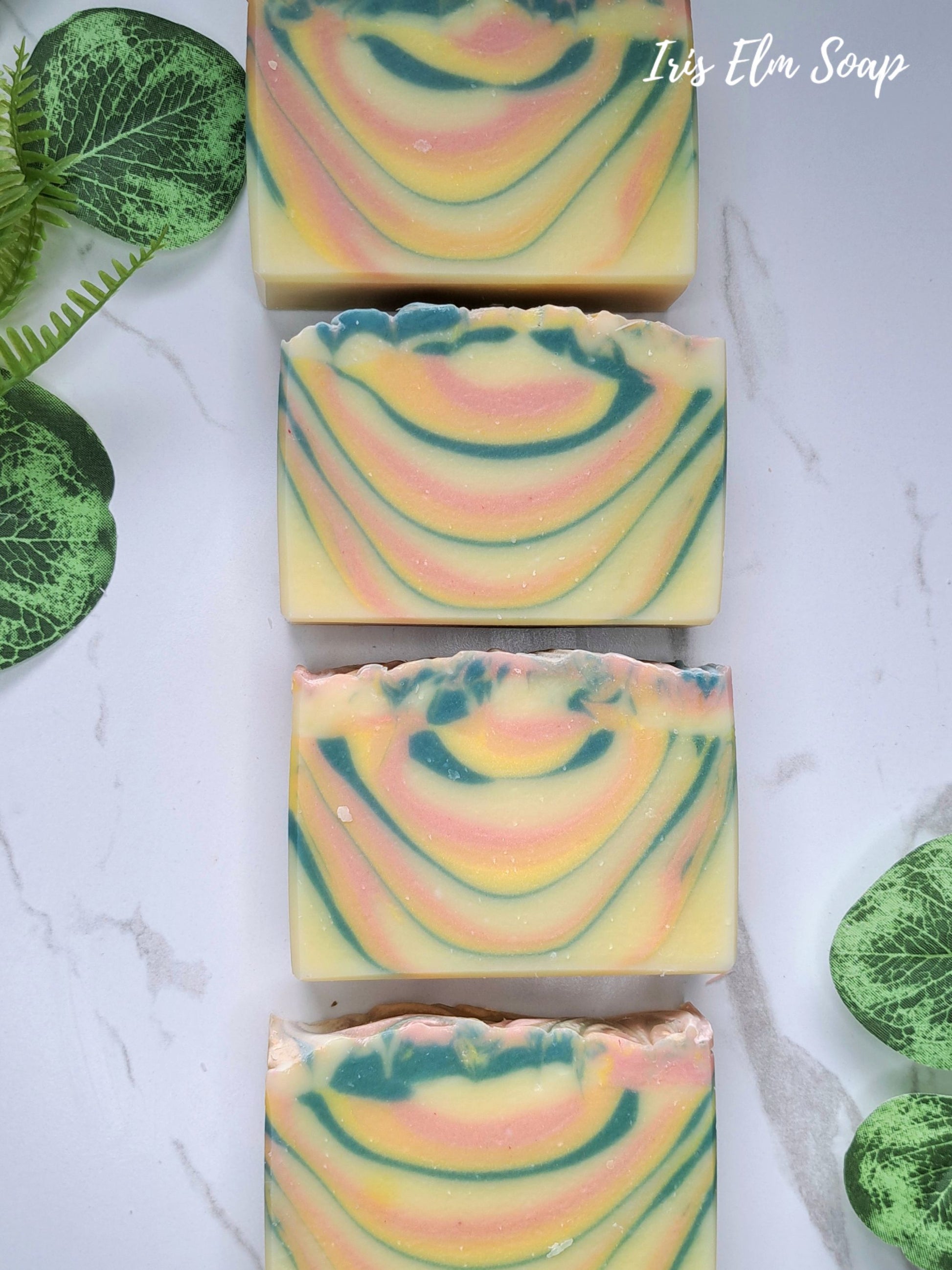 Four rectangular bars of orange patchouli artisanal soap with a marbled swirl pattern of yellow, pink, orange, and teal, arranged vertically on a white marble surface with green decorative leaves.