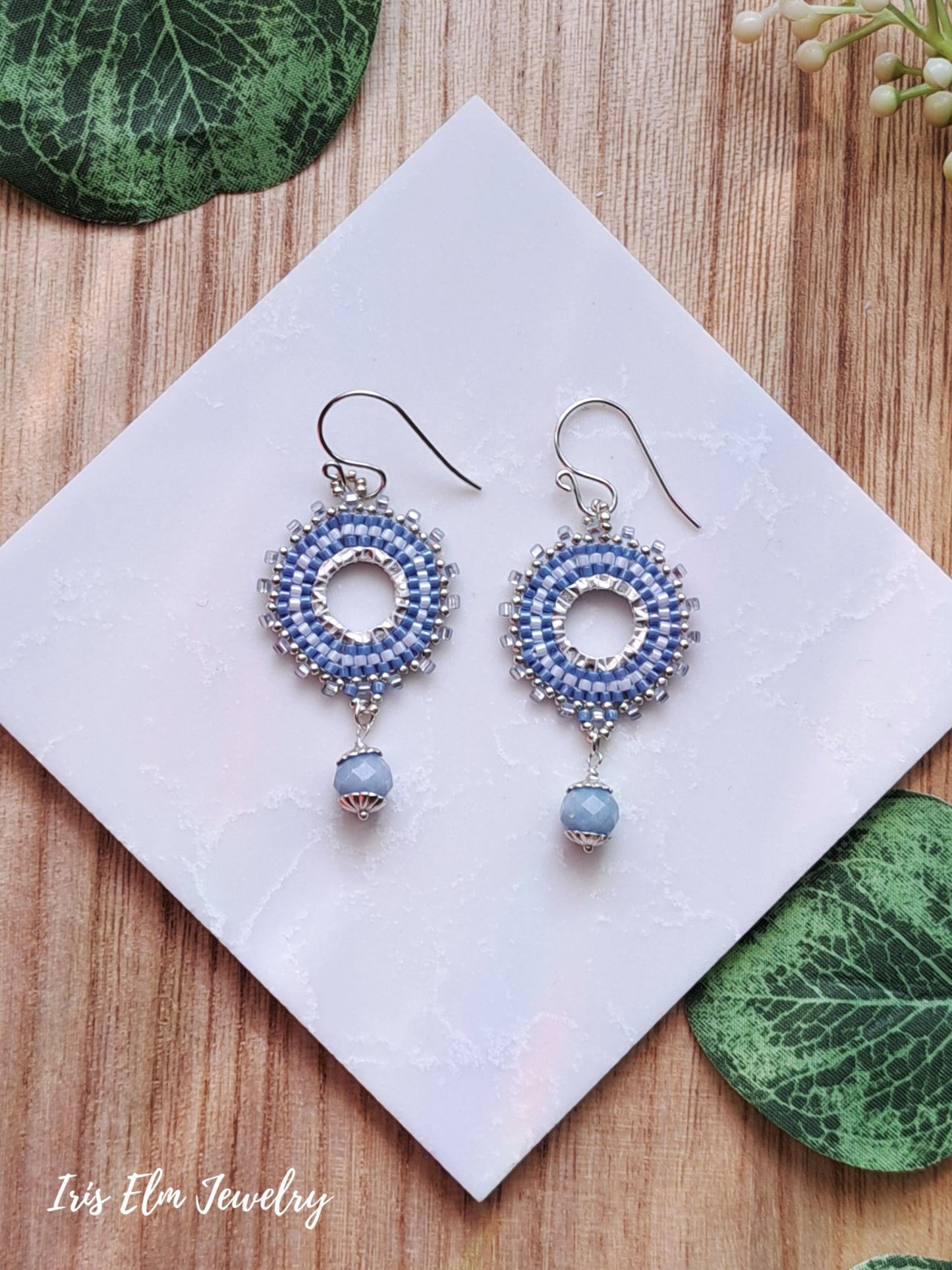 Pair of blue beaded hoop earrings with angelite stone dangles on a white card with green leaves on a wooden surface