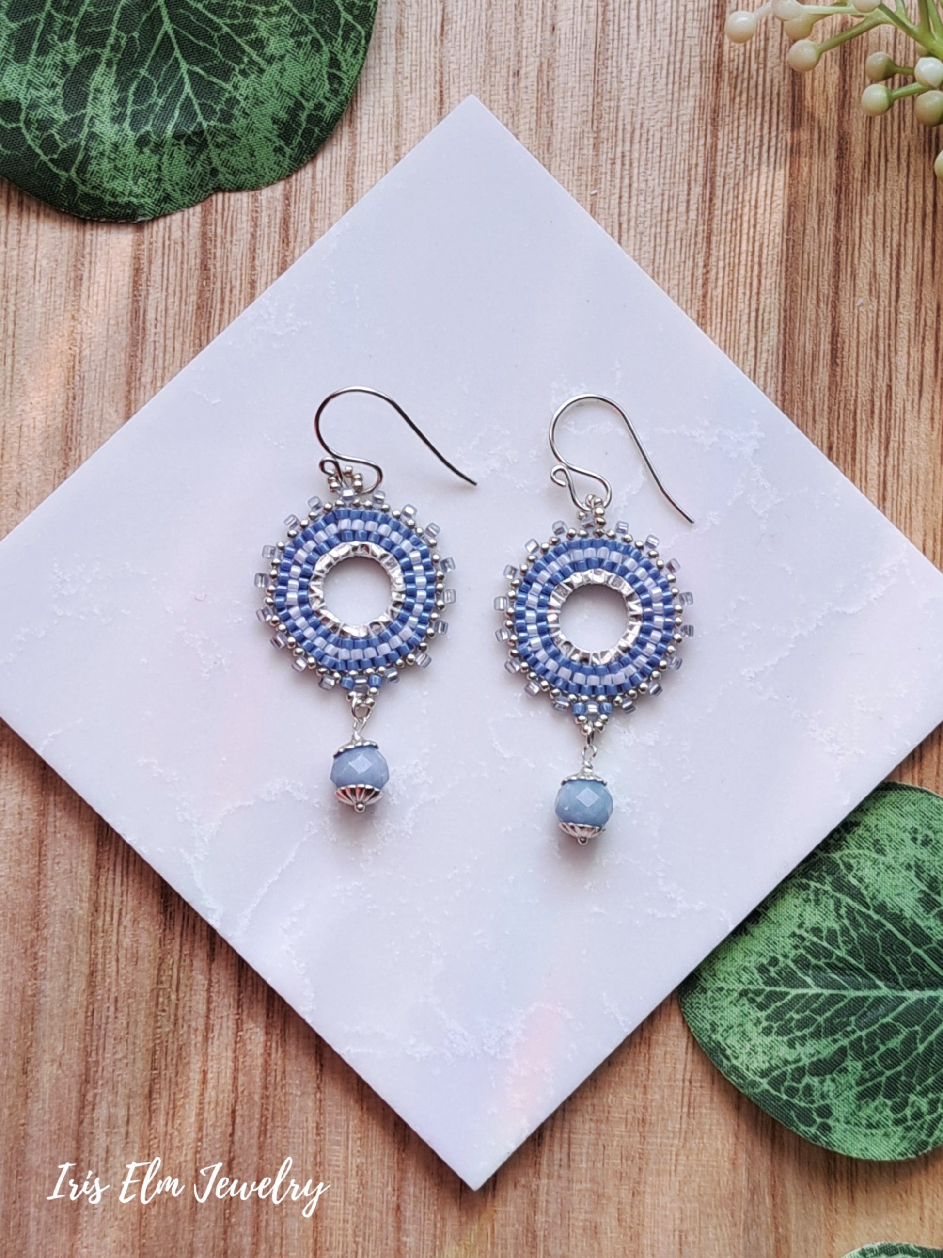 Pair of blue beaded hoop earrings with angelite stone dangles on a white card with green leaves on a wooden surface