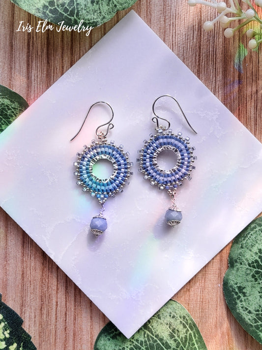 Pair of beaded blue seed bead and angelite hoop earrings on a white napkin with 'Iris Elm Jewelry' branding.