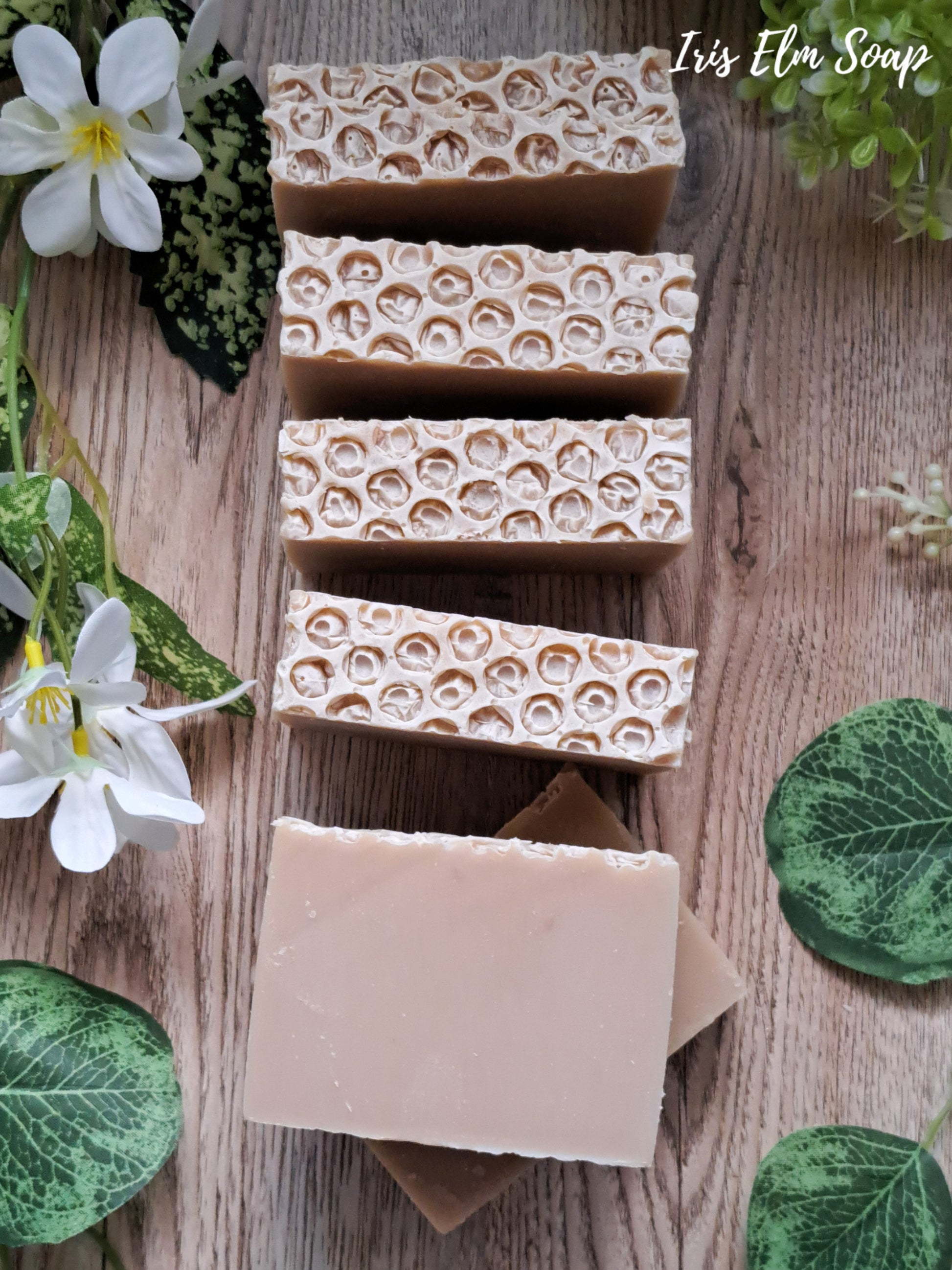 Beige oatmeal honey handmade soap bars with honeycomb pattern on a wooden surface with green leaves and flowers.