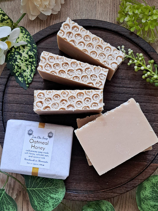Three bars of oatmeal honey handmade soap with a packaging label on a wooden surface with greenery.