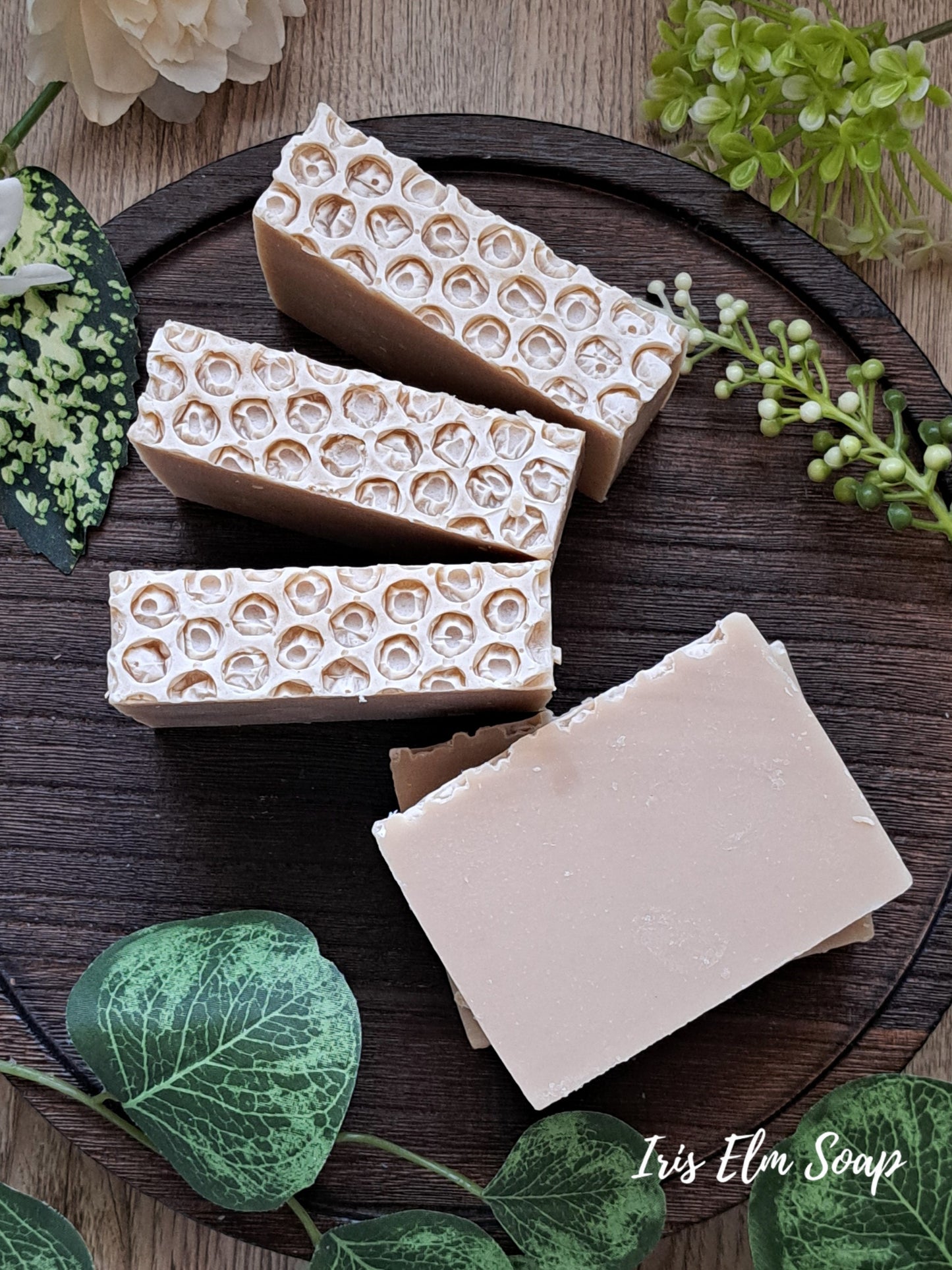 Three bars of oatmeal honey handmade soap with a honeycomb pattern on a wooden board, surrounded by greenery.