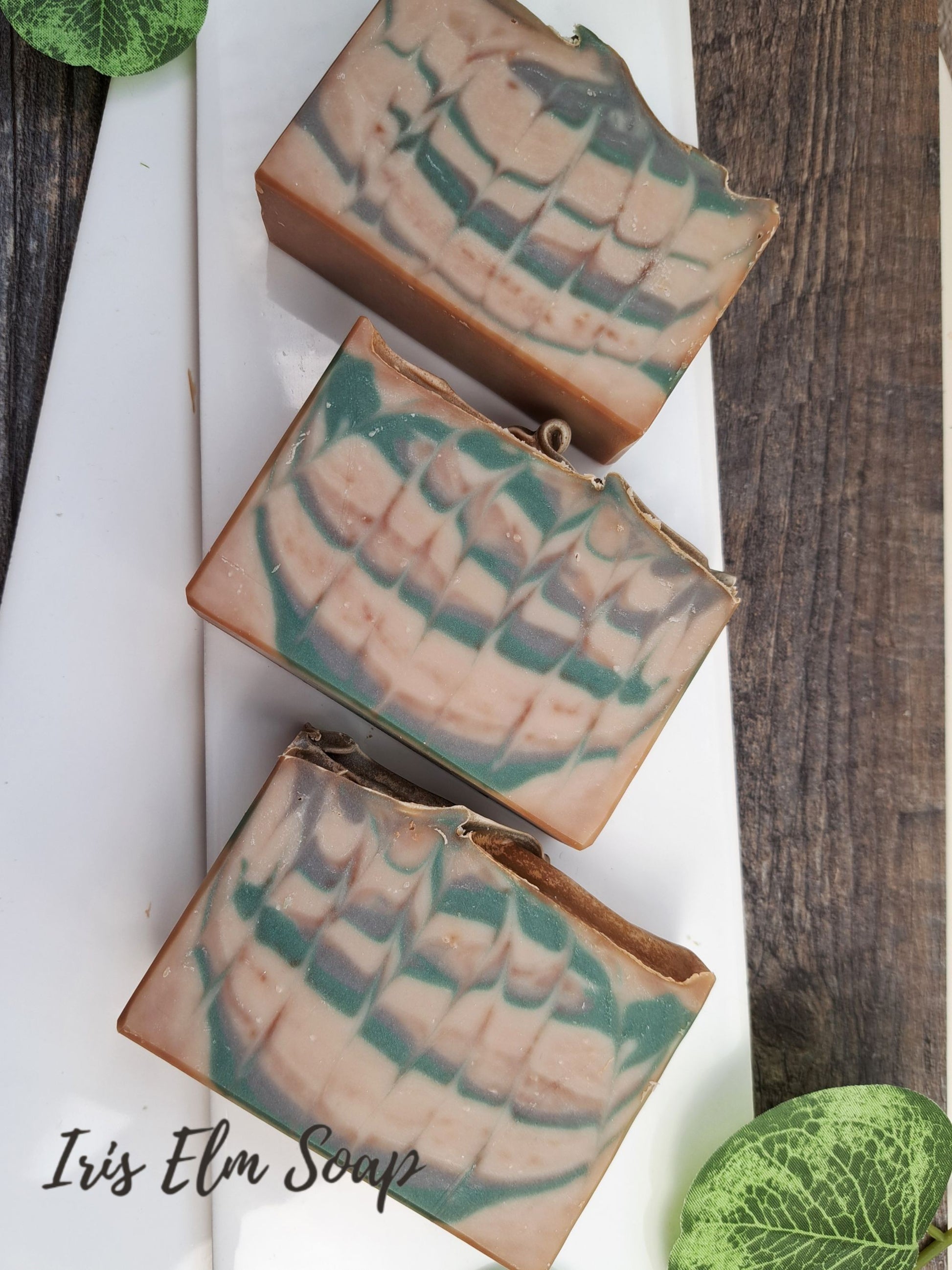 Three bars of Iris Elm Soap on a wooden surface with green leaves.