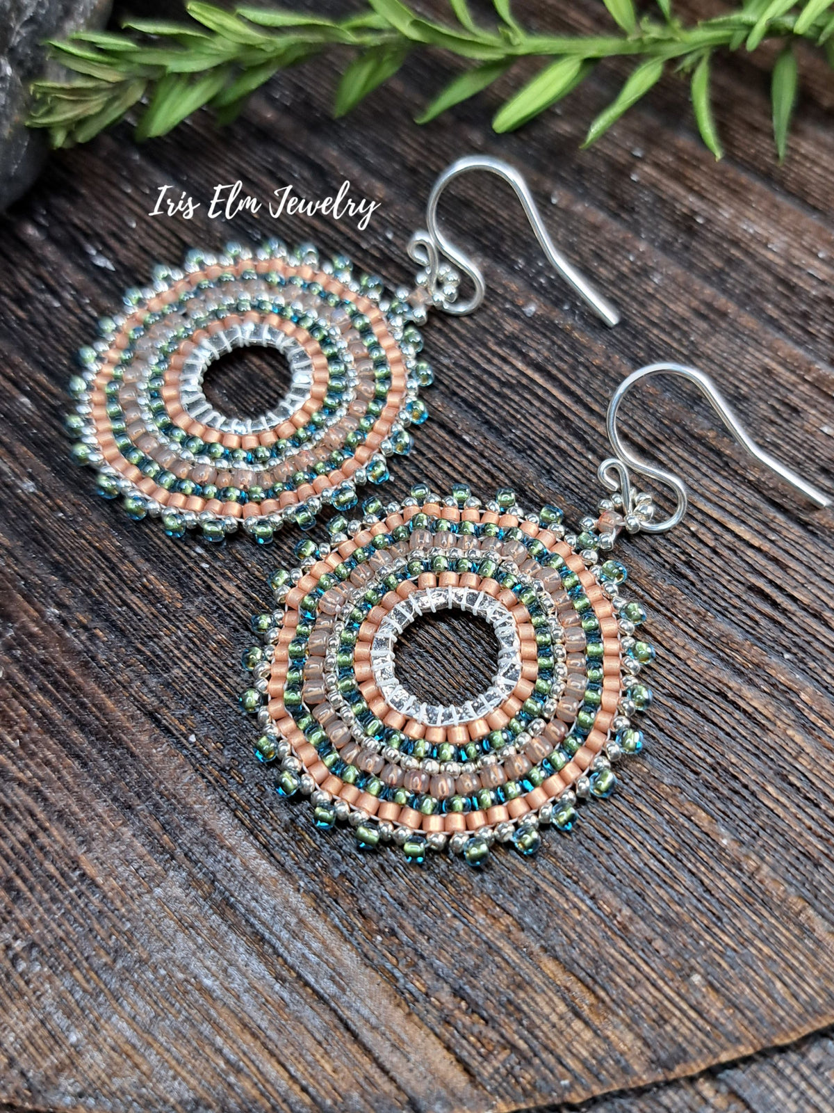 A pair of handmade beaded dangle earrings with silver fishhook wires, featuring concentric circles of peach, green, and silver beads. The earrings are displayed on a dark wood surface with green leaves in the background, and the brand name 'Iris Elm Jewelry' is visible as an overlay.