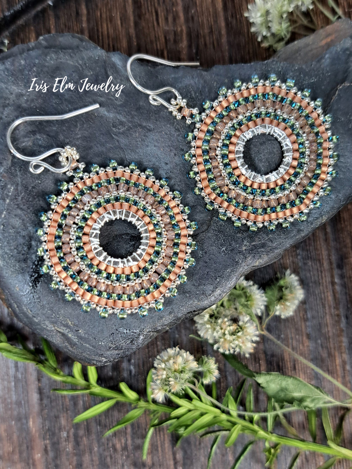 A pair of handmade beaded drop earrings by Iris Elm Jewelry, featuring concentric circles of peach, green, and silver beads. The earrings are displayed on a dark slate stone with green sprigs and small white flowers on a rustic wooden surface.