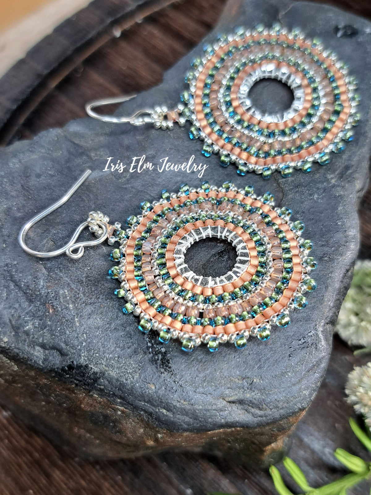 A pair of handmade beaded dangle earrings with silver fishhook wires, featuring concentric circles of peach, teal, and silver beads. The earrings are displayed on a dark textured stone with a wooden surface in the background.