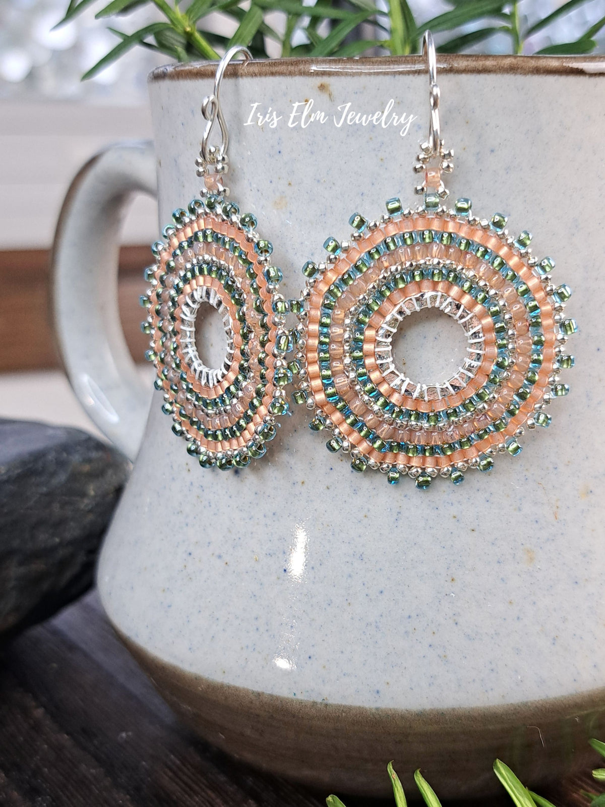 A pair of handmade spiral beaded earrings with peach, green, and silver beads, hanging from silver fishhook wires. The earrings are displayed on a speckled ceramic mug with green foliage in the background.