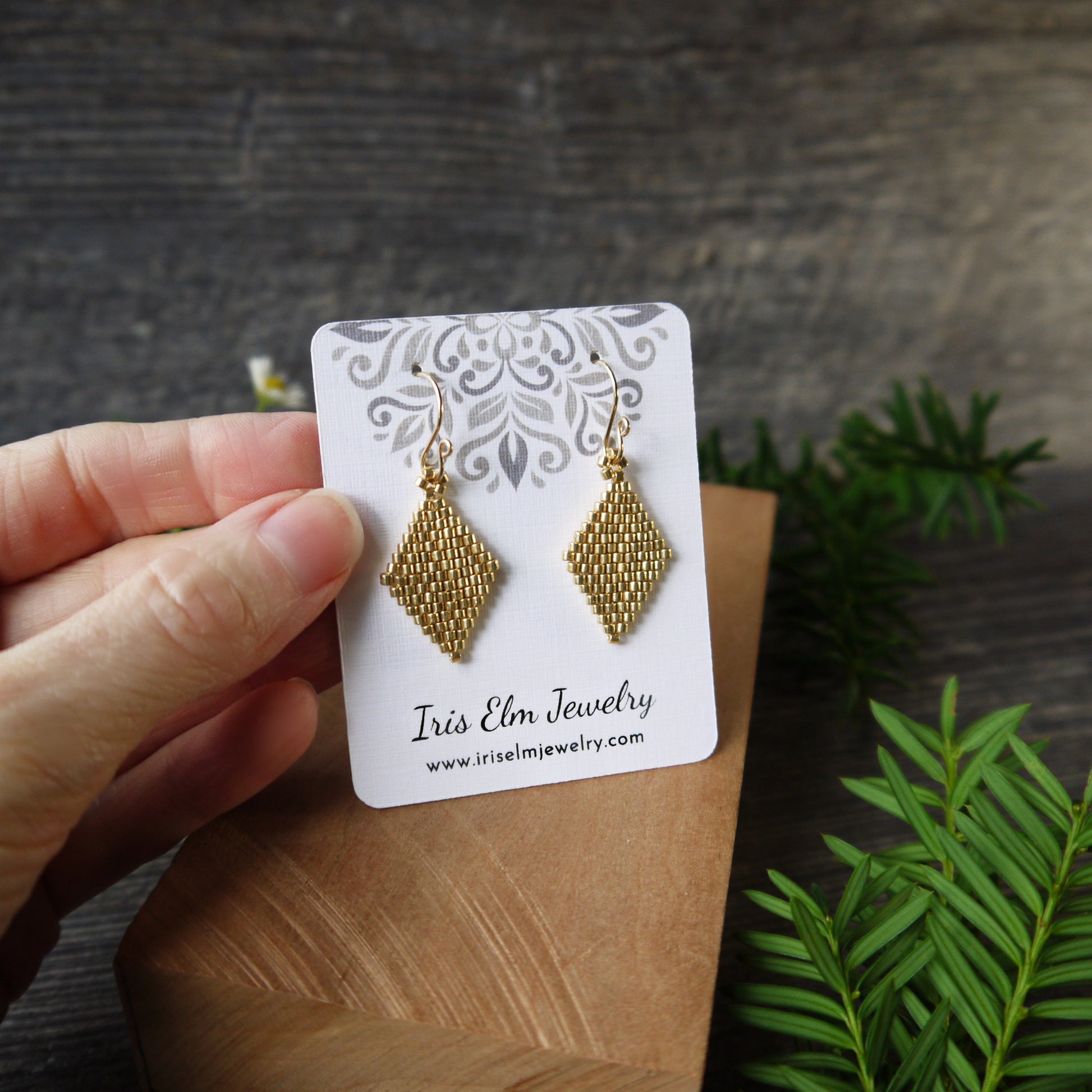 Gold diamond shape seed bead handmade earrings on a 'Iris Elm Jewelry' card held by a hand with a natural background