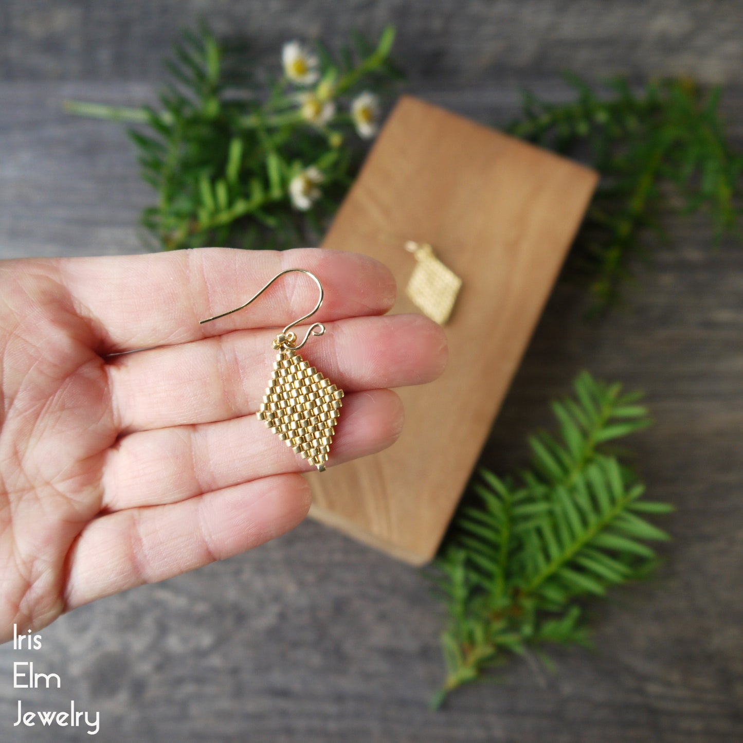 Gold beaded seed bead diamond earring held in a hand with a blurred background of greenery and a wooden block.