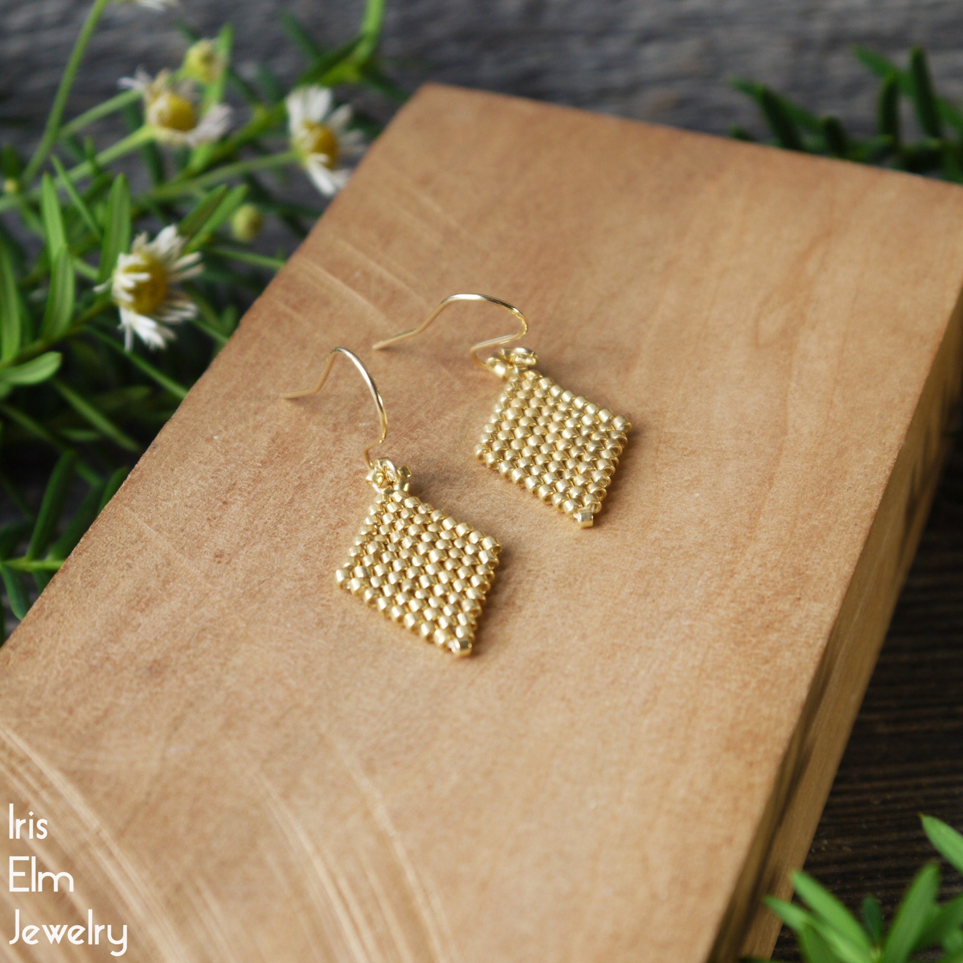 Gold diamond shape beaded earrings on a wooden block with flowers in the background, featuring 'Iris Elm Jewelry'.