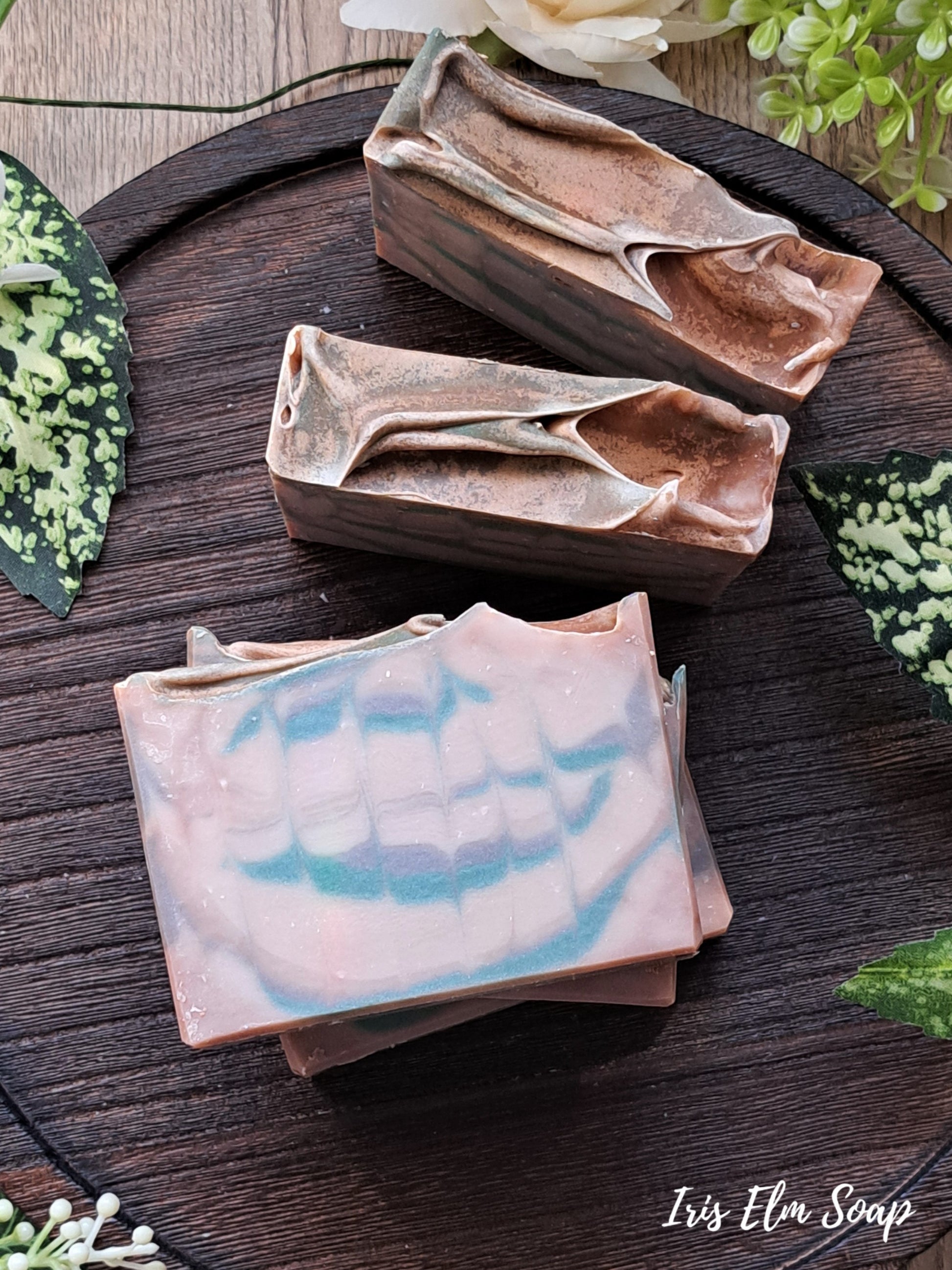 Three soap Minnesota north woods handmade bars with swirl design on a wooden surface, surrounded by greenery.