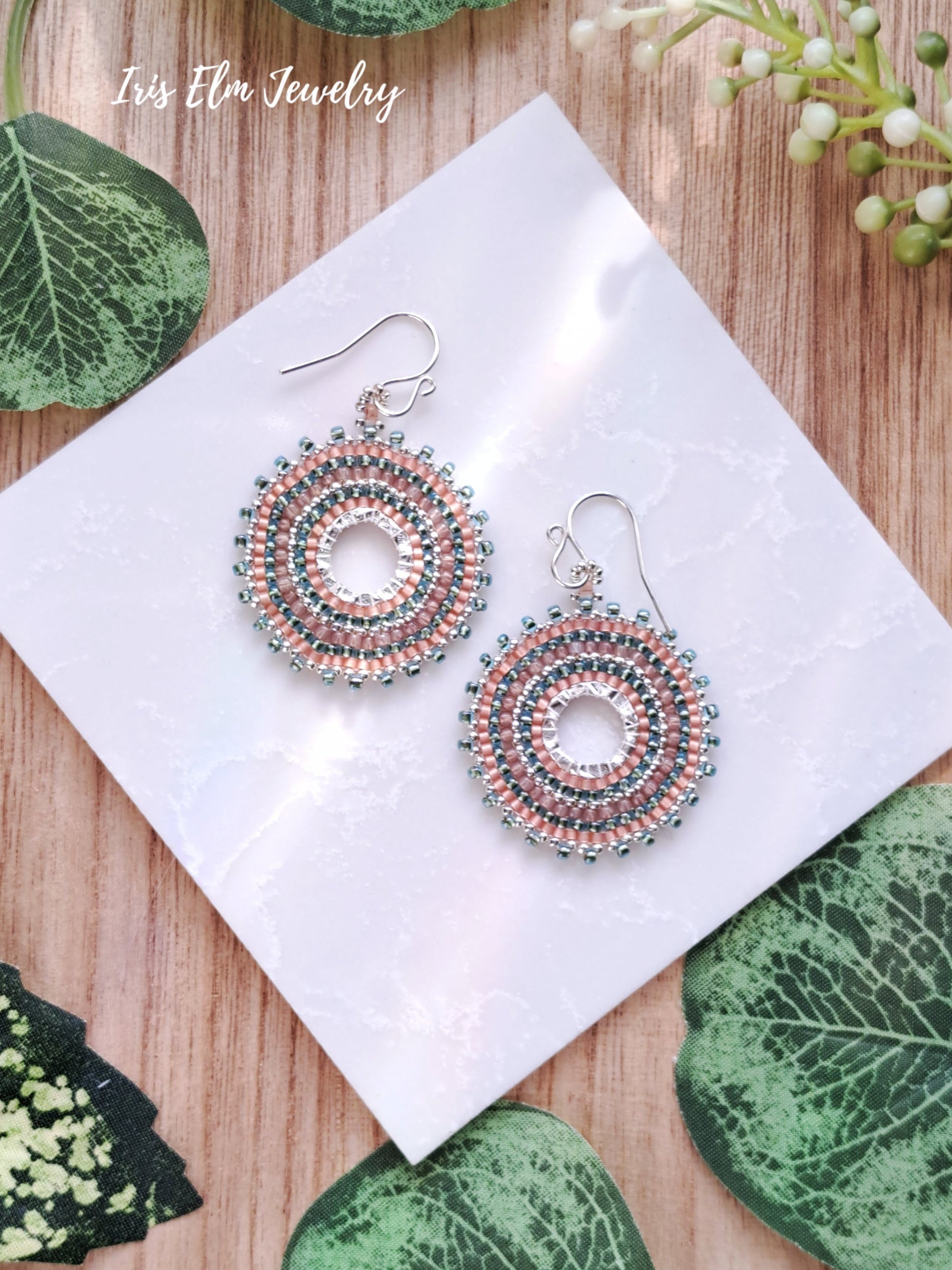 Decorative peach and aqua seed bead beadwork glass hoop earrings on a white napkin with green leaves in the background, branded 'Iris Elm Jewelry'.
