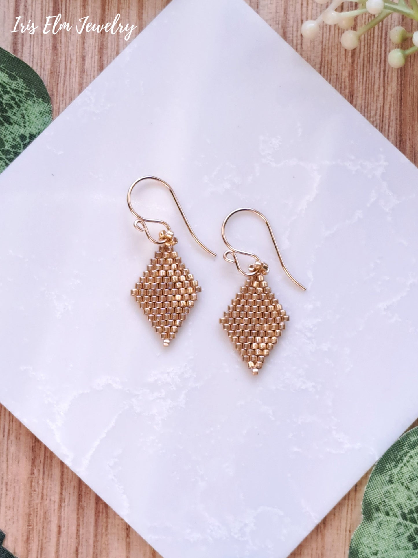 Gold beaded seed bead diamond shape small earrings on a white cloth with 'Iris Elm Jewelry' branding.
