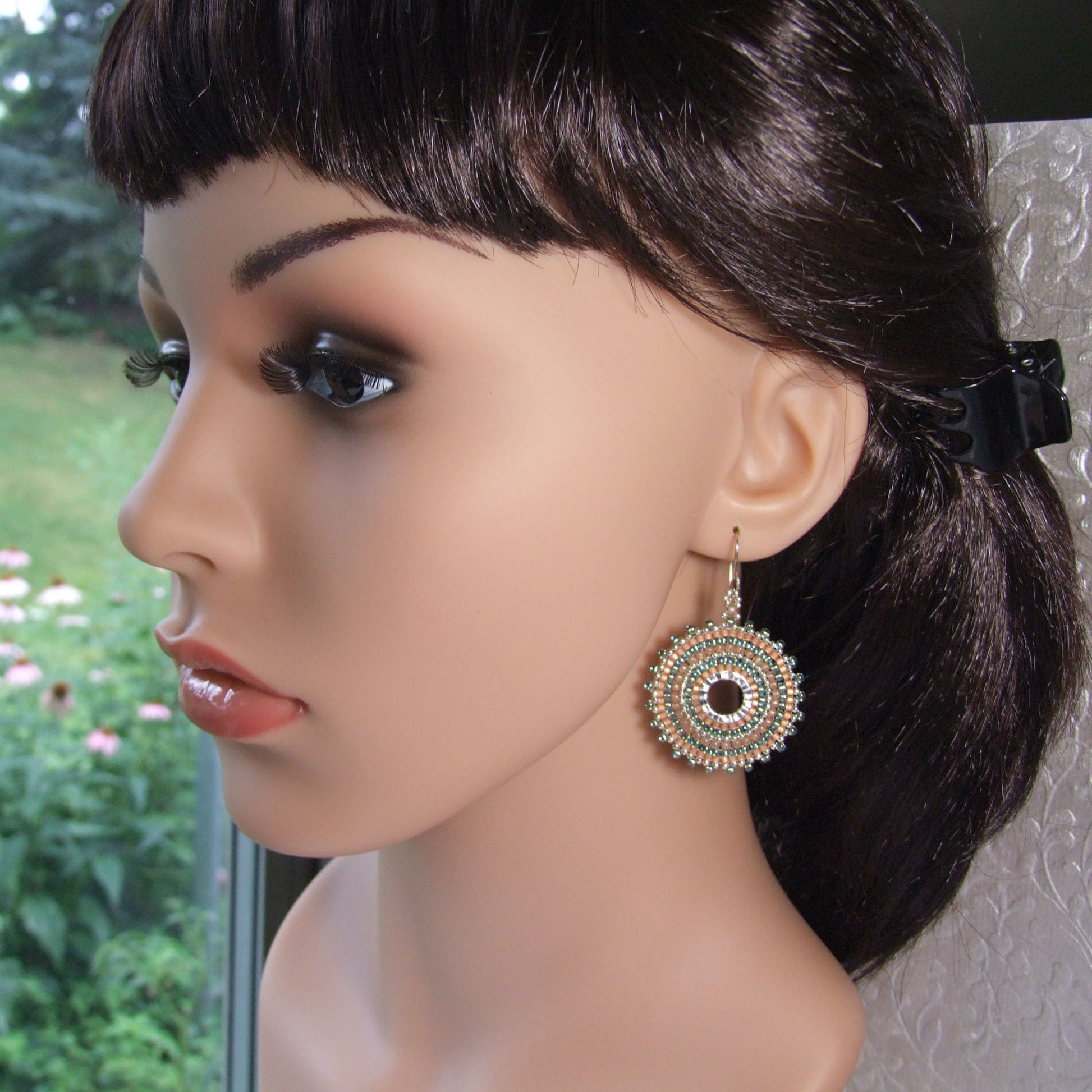 Mannequin head with a decorative peach and aqua woven seed bead hoop earring on a blurred outdoor background
