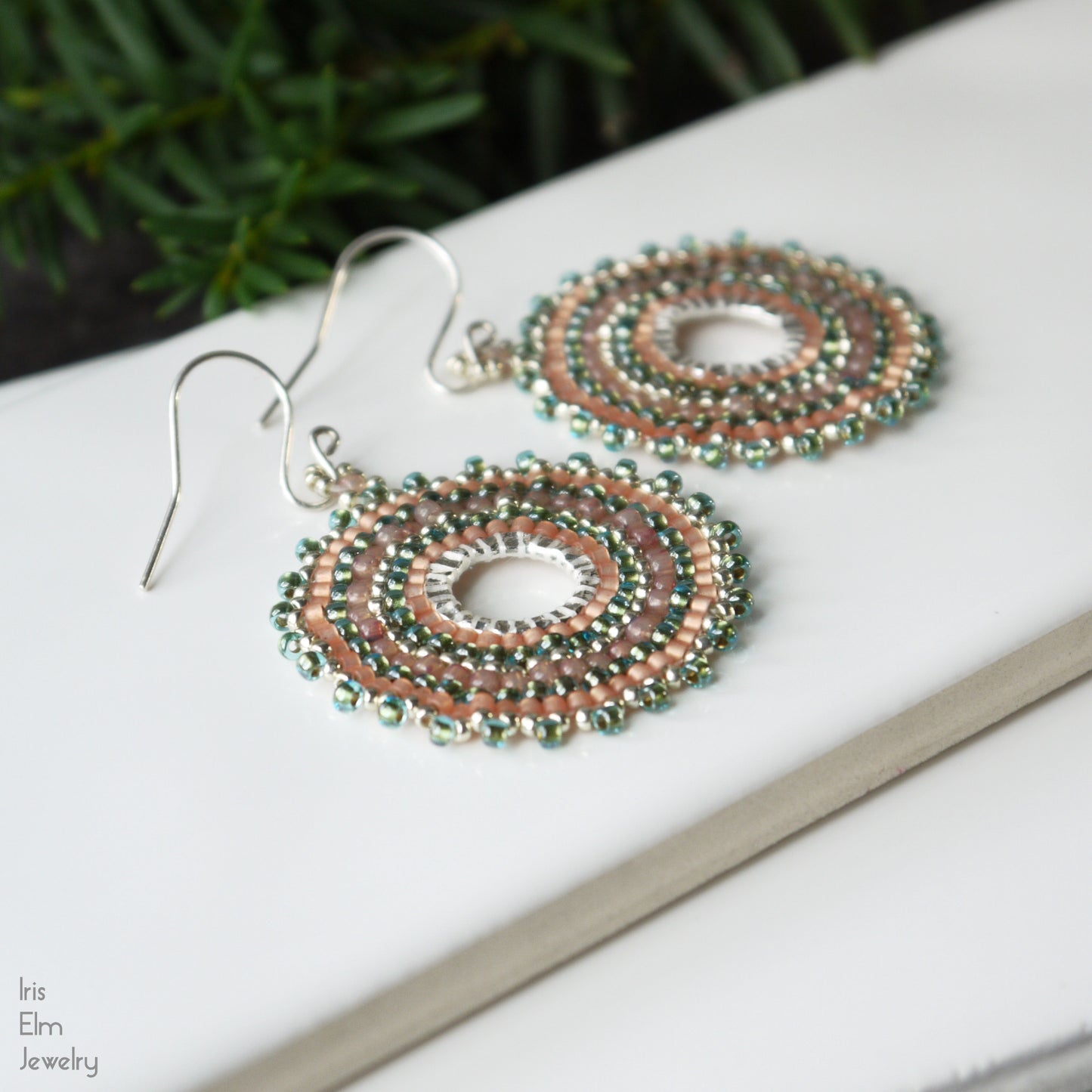 Pair of beaded aqua green, silver, and peach seed bead earrings on a white surface with greenery in the background
