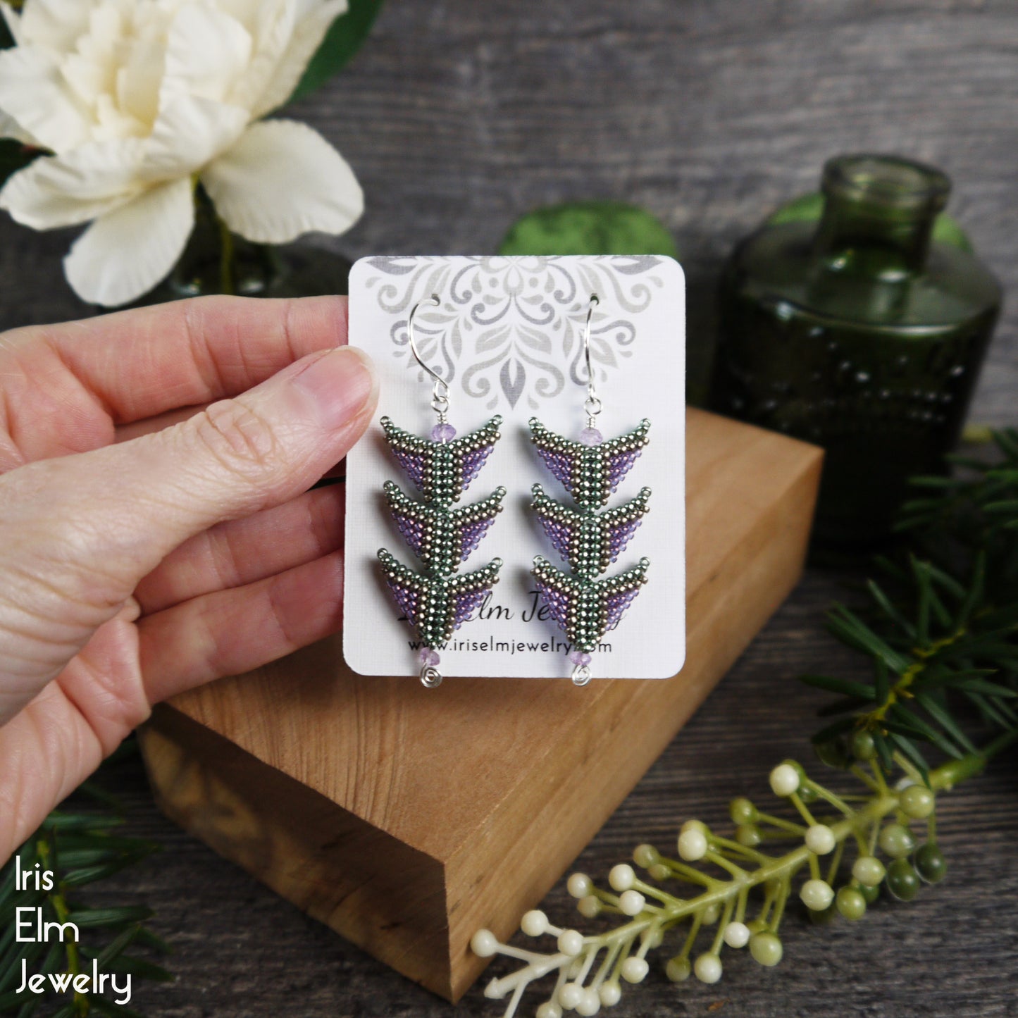 Hand holding a card with amethyst and seed bead purple and green triangle earrings on a wooden block with decorative elements.