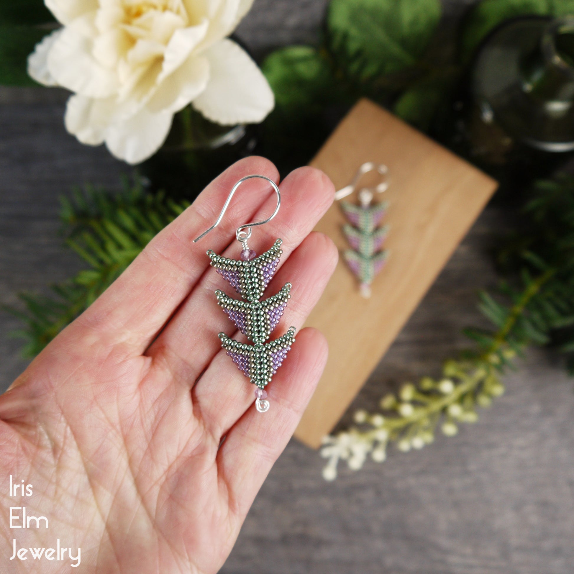Hand holding a beadwork glass seed bead triangle tiered purple and green earring with floral and wooden background