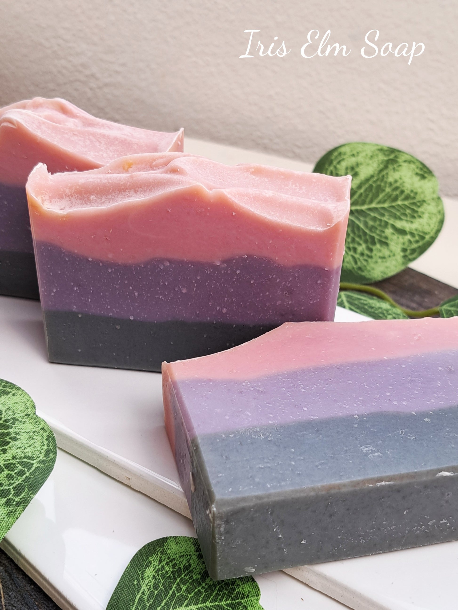 Three layered handmade soap bars with pink, purple, and black layers on a white surface with green leaves.