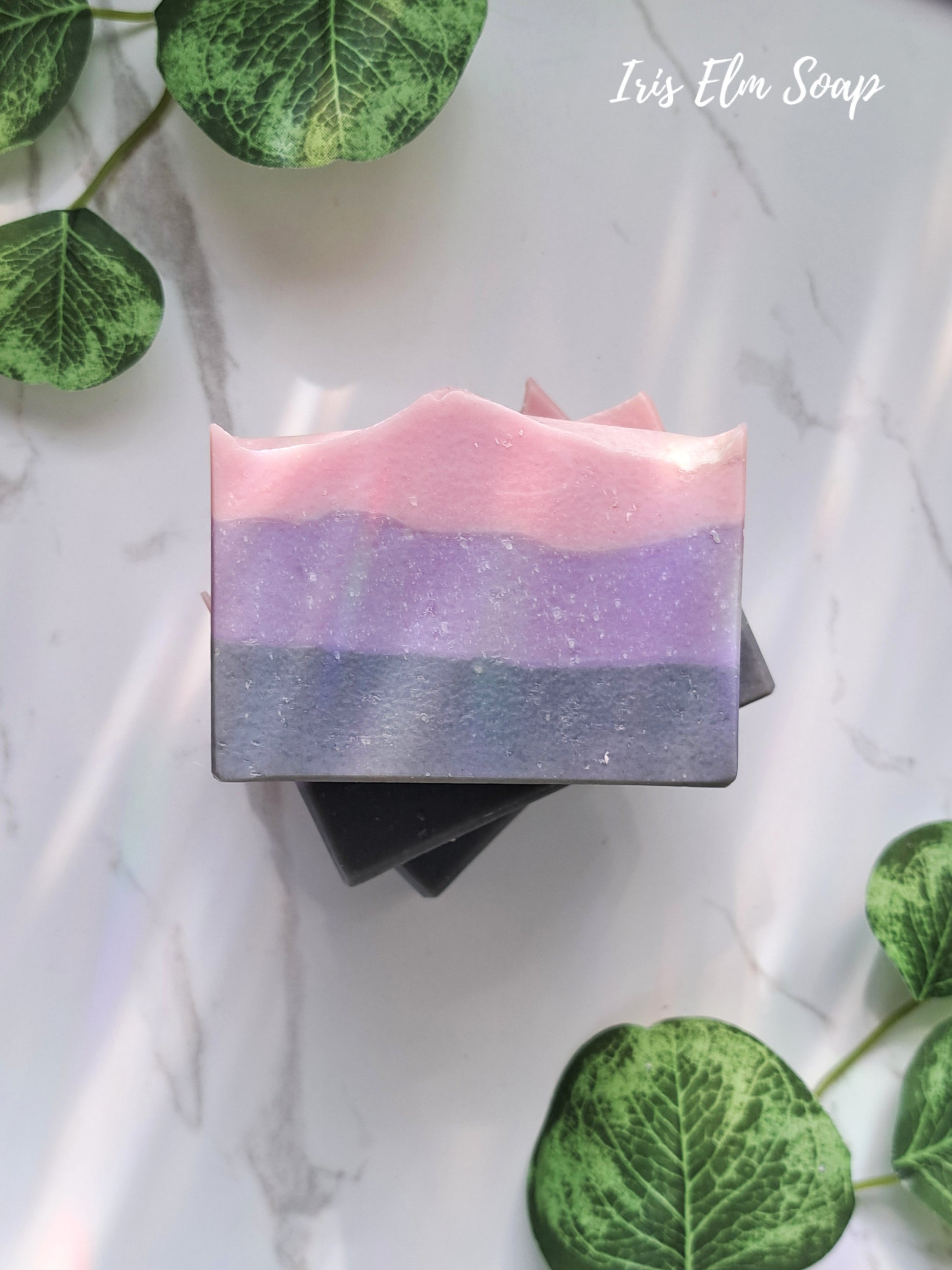 Three layered soap moonlit peony handmade bar with pink, purple, and gray on a marble surface with green leaves.