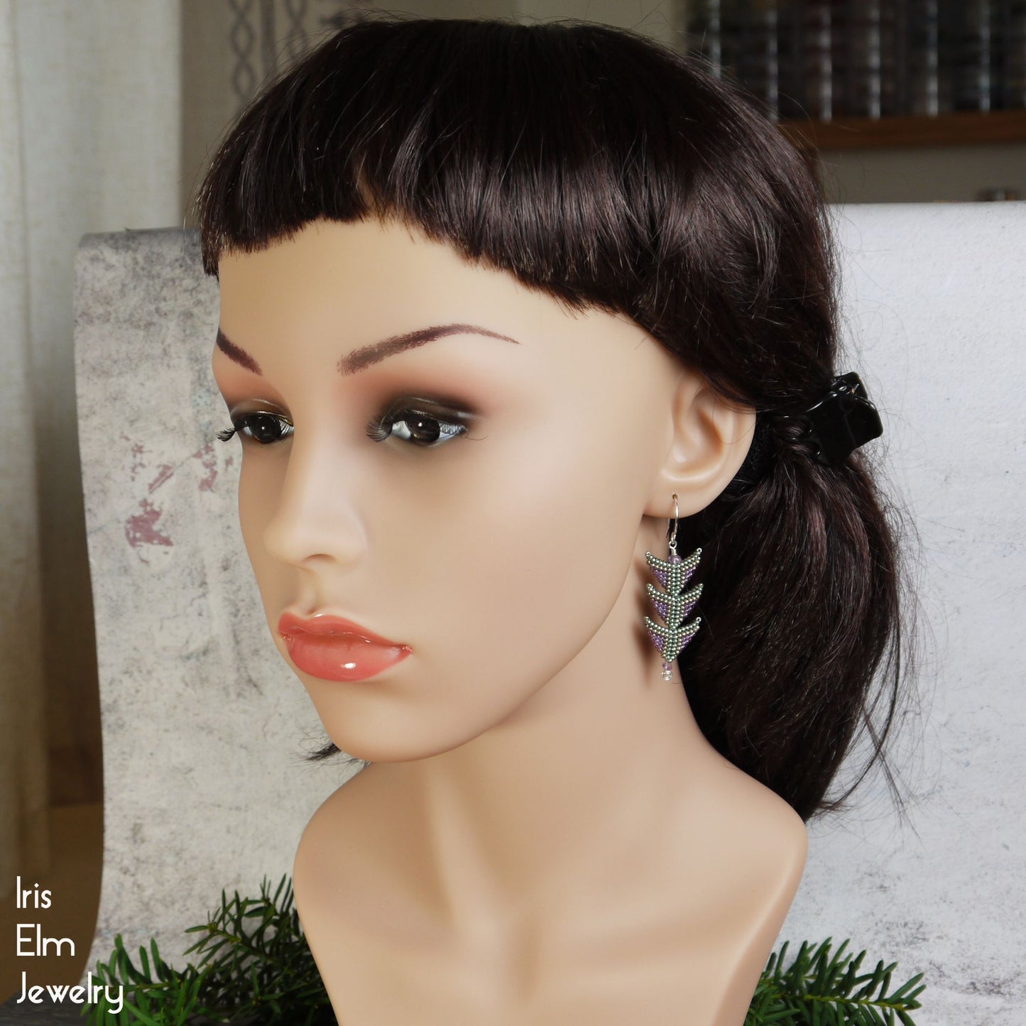 Mannequin head with dark hair styled in a ponytail, wearing tiered triangle earrings with a blurred background.