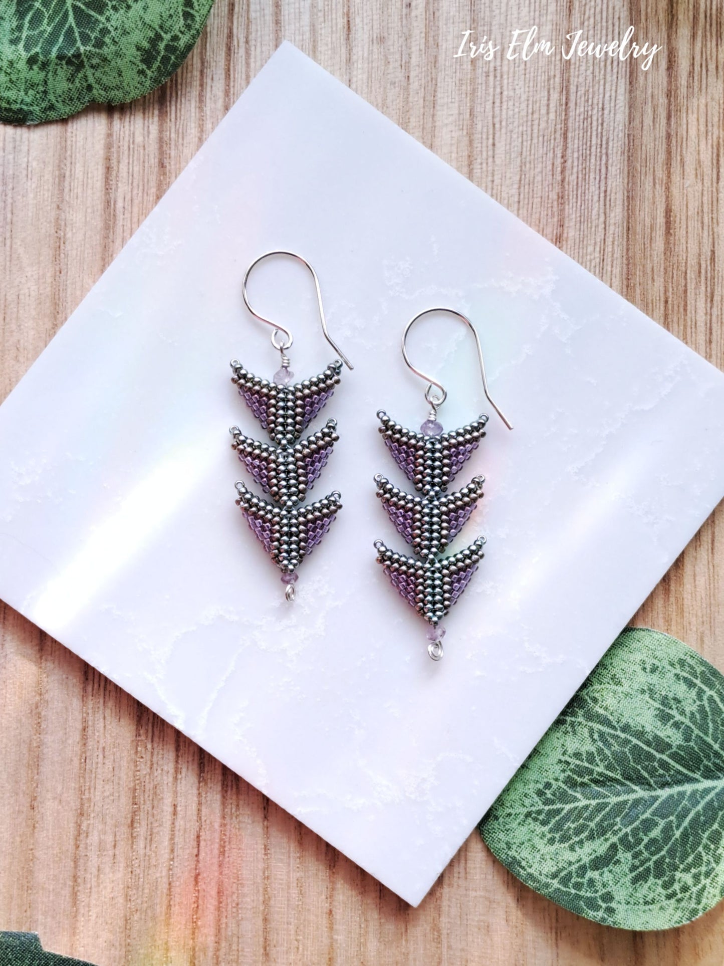 Pair of beaded purple green triangle seed bead amethyst earrings on a white card with 'Iris Elm Jewelry' branding, placed on a wooden surface with green leaves.