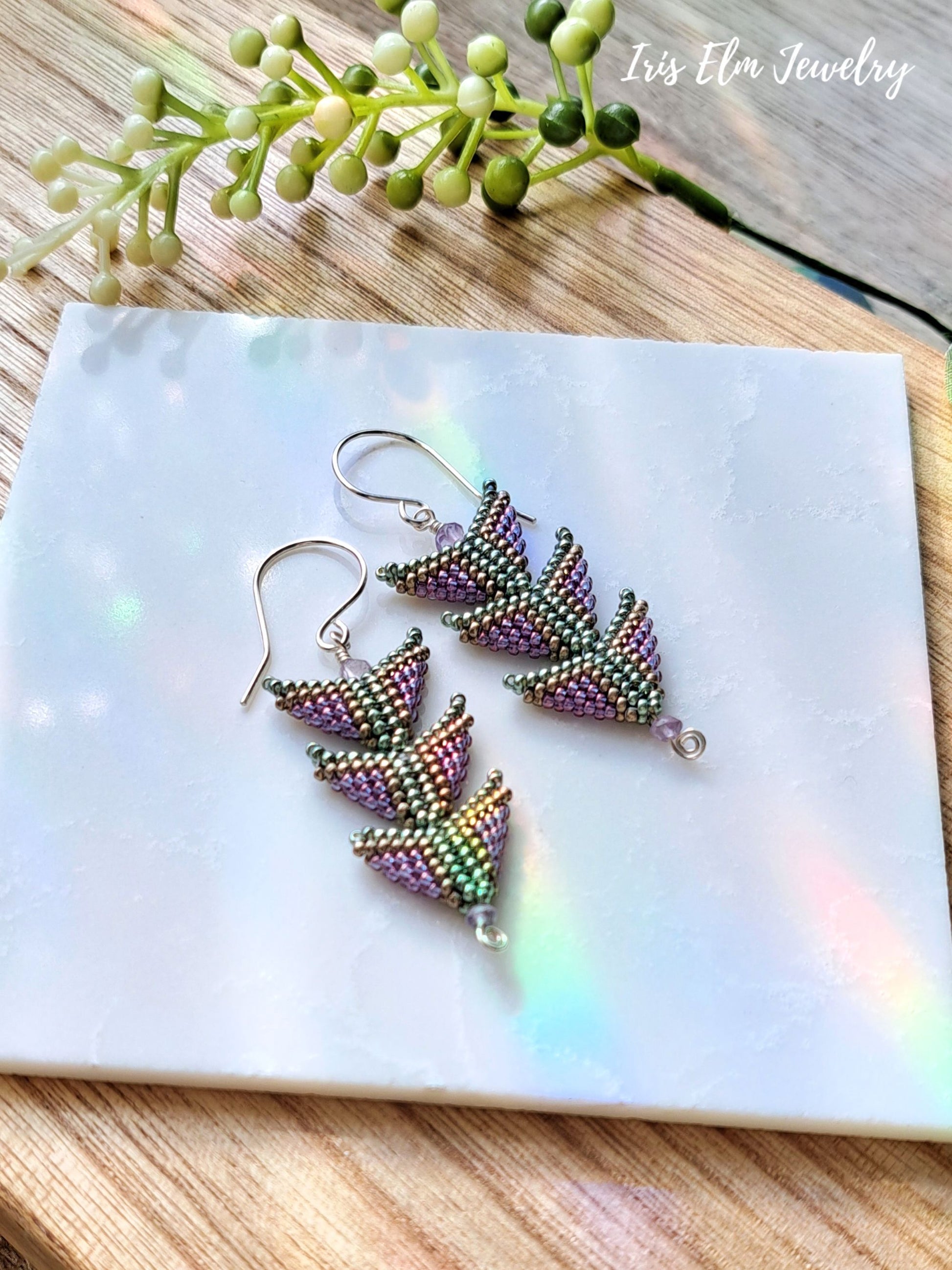 Pair of beaded purple green triangle seed bead amethyst sterling silver earrings on a white card with 'Iris Elm Jewelry' branding, placed on a wooden surface with green leaves.