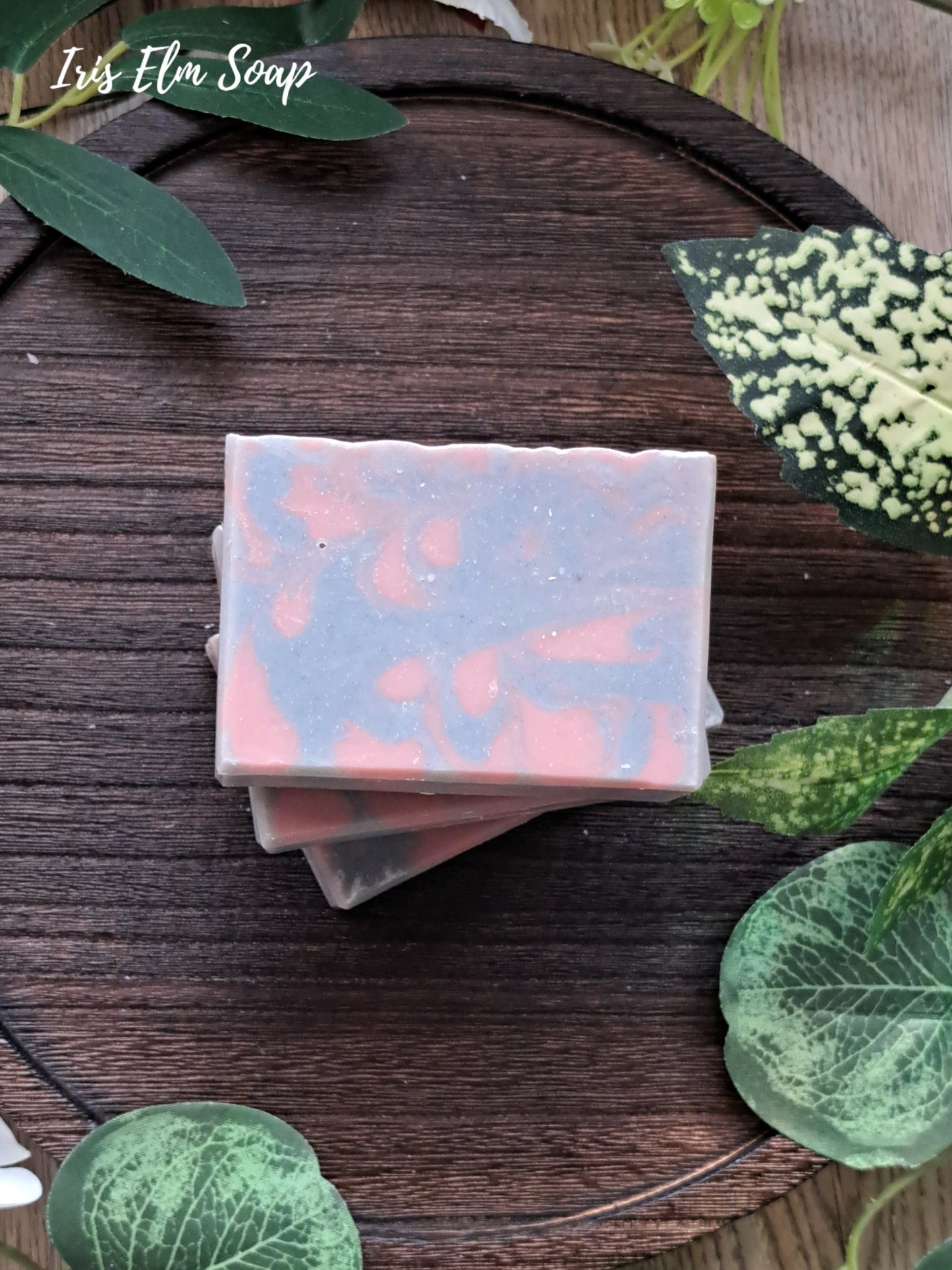 Stack of lavender essential oil handmade soap bars with rose clay and indigo swirl on a wooden surface with green leaves.
