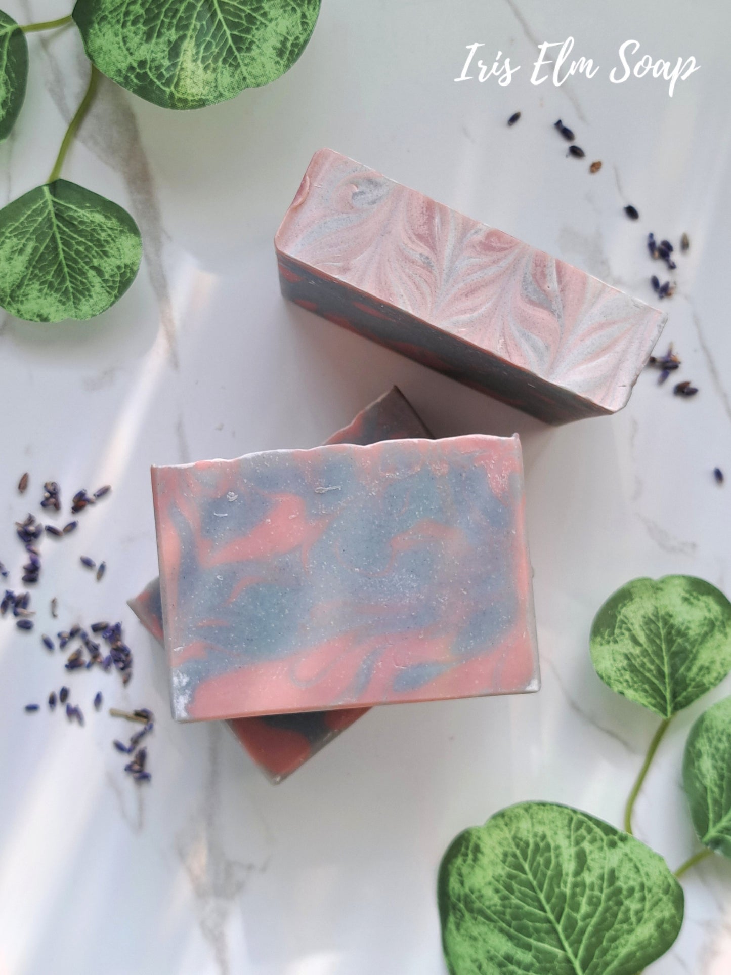 Three lavender handmade soap bars with a marbled design on a white surface with green leaves and lavender flowers.