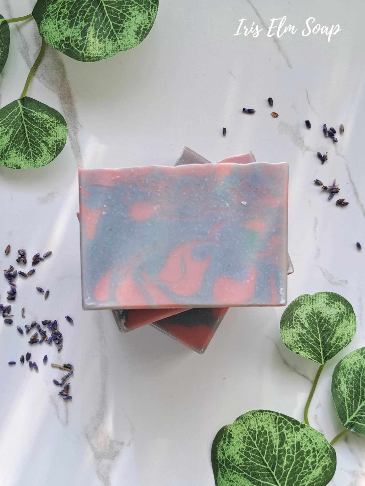 Stack of colorful lavender essential oil handmade soap bars with green leaves and lavender flowers on a white background, featuring 'Iris Elm Soap'.