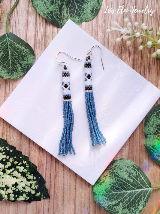 Blue tassel earrings on a white square with green leaf decorations on a wooden surface.